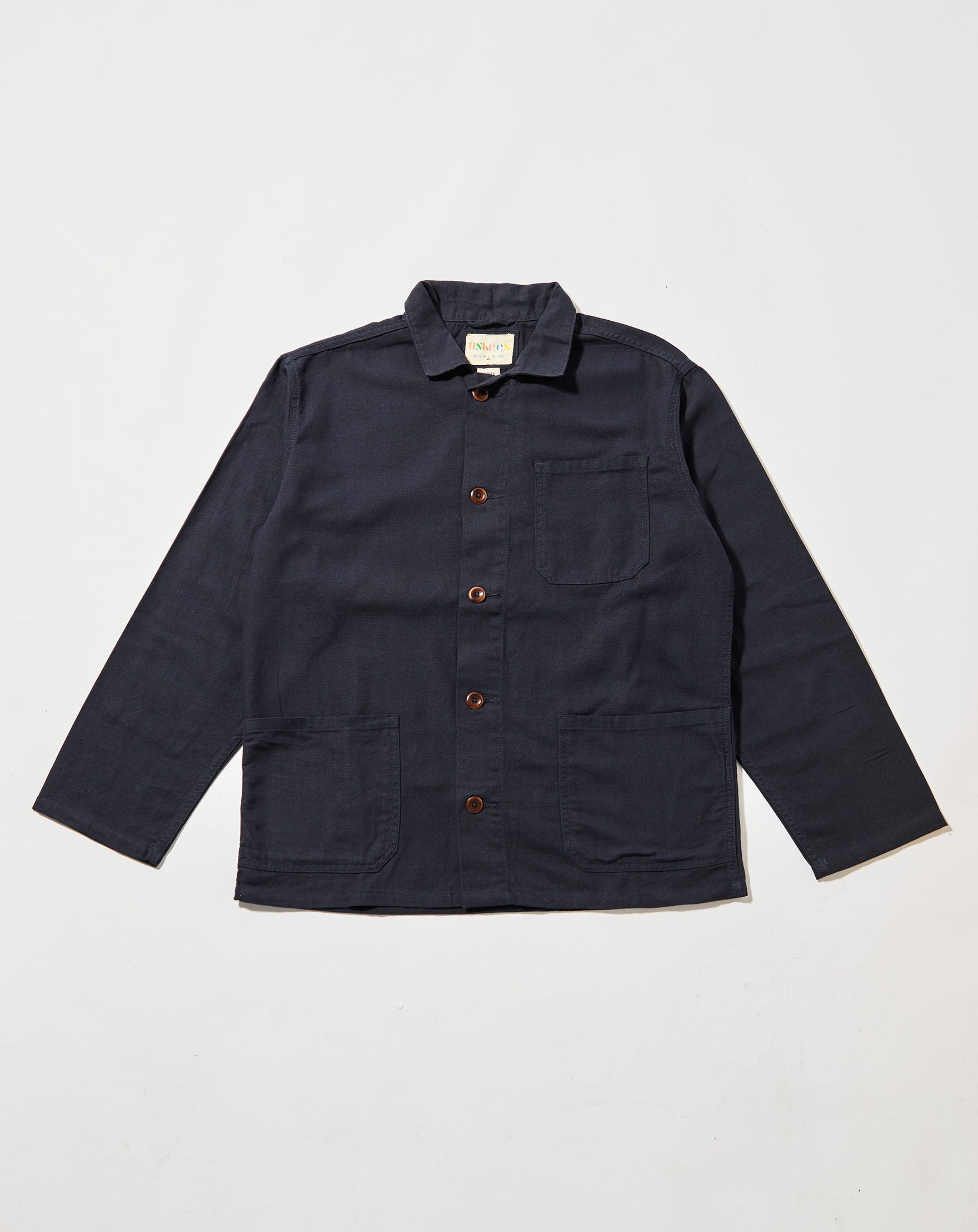 Front flat shot of midnight blue, buttoned organic cotton overshirt. Clear view of chest and hip pockets, corozo buttons and Uskees branding label.  Zero waste production.