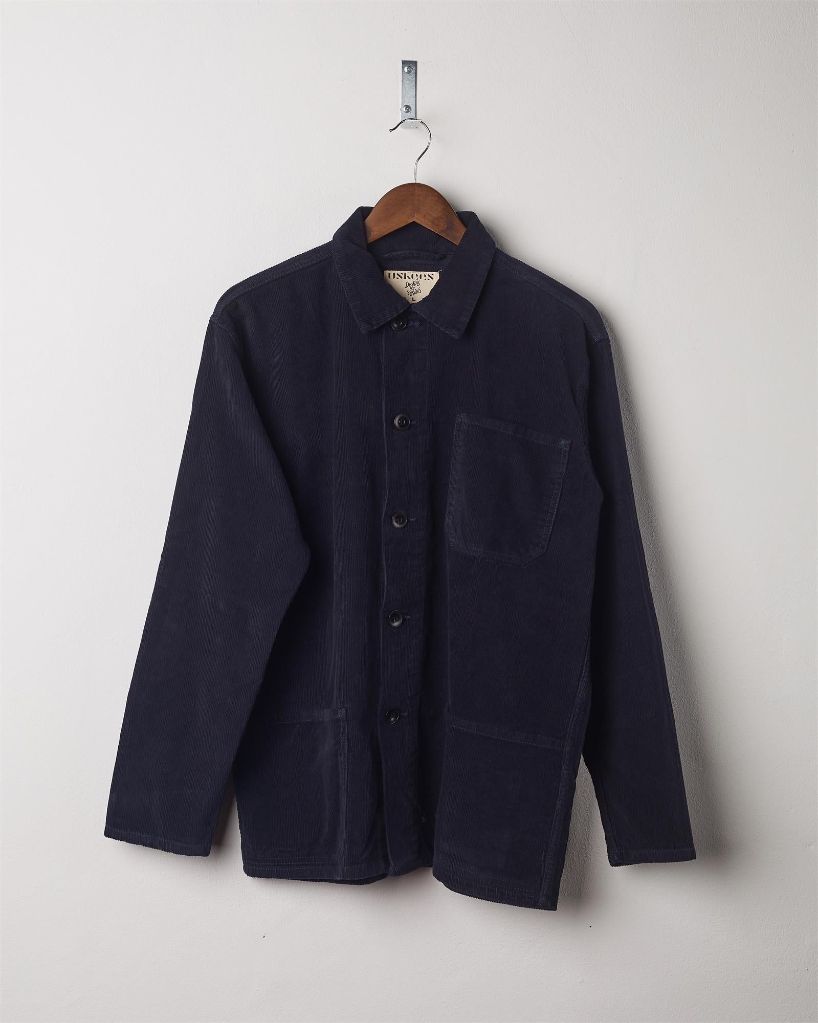 Midnight blue coloured, buttoned corduroy overshirt from Uskees presented on hanger. Clear view of breast and hip pocket and corozo buttons.