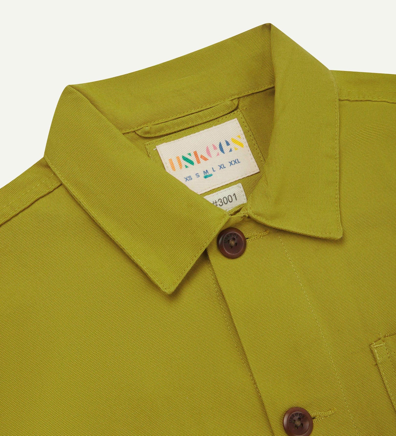 Close-up top-half view of #3001, yellow-green pear-coloured organic cotton drill overshirt. With focus on collar, Uskees brand label and corozo buttons.