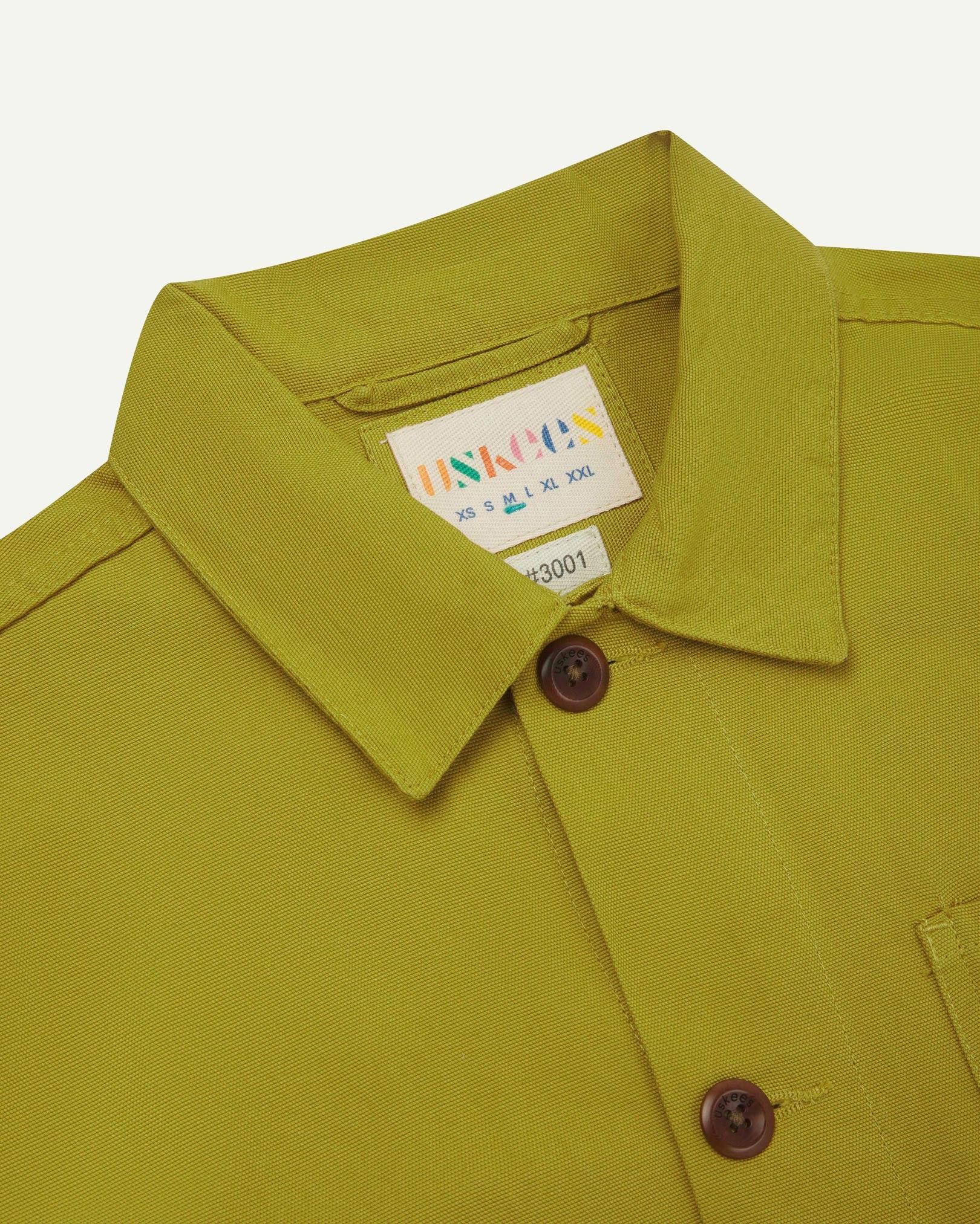 Close-up top-half view of #3001, yellow-green pear-coloured organic cotton drill overshirt. With focus on collar, Uskees brand label and corozo buttons.