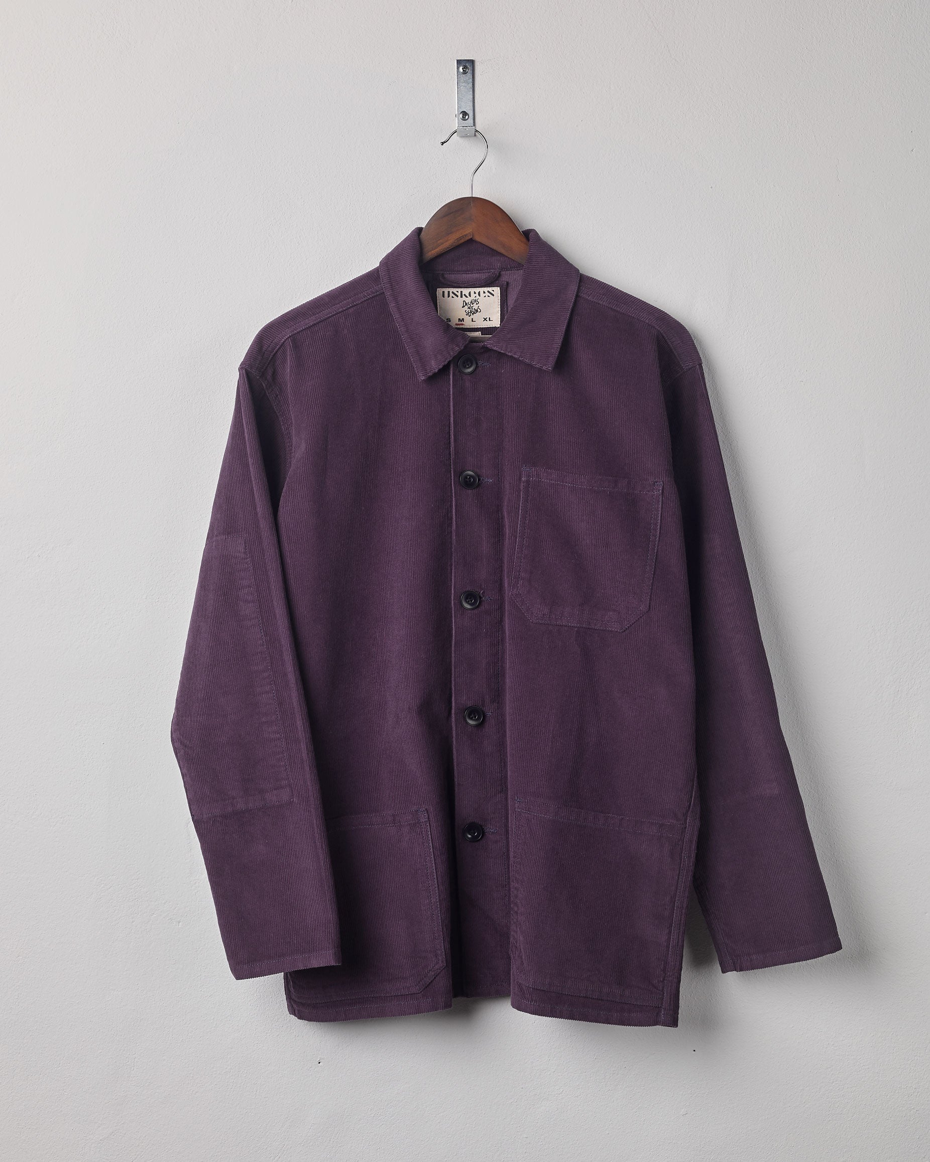 Plum coloured, buttoned corduroy overshirt from Uskees presented on hanger. Clear view of breast and hip pocket and corozo buttons.