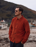 Man wearing an Uskees rust red/orange cord overshirt and sunglasses on a beach with mountains in the background