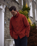 Man wearing an Uskees rust corduroy shirt-jacket standing in front of a stone building with greenery.