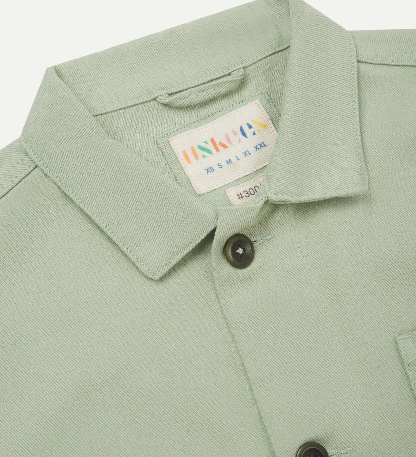 Close-up top-half view of #3001 silver-green organic cotton drill overshirt. With focus on collar, Uskees brand label and corozo buttons.