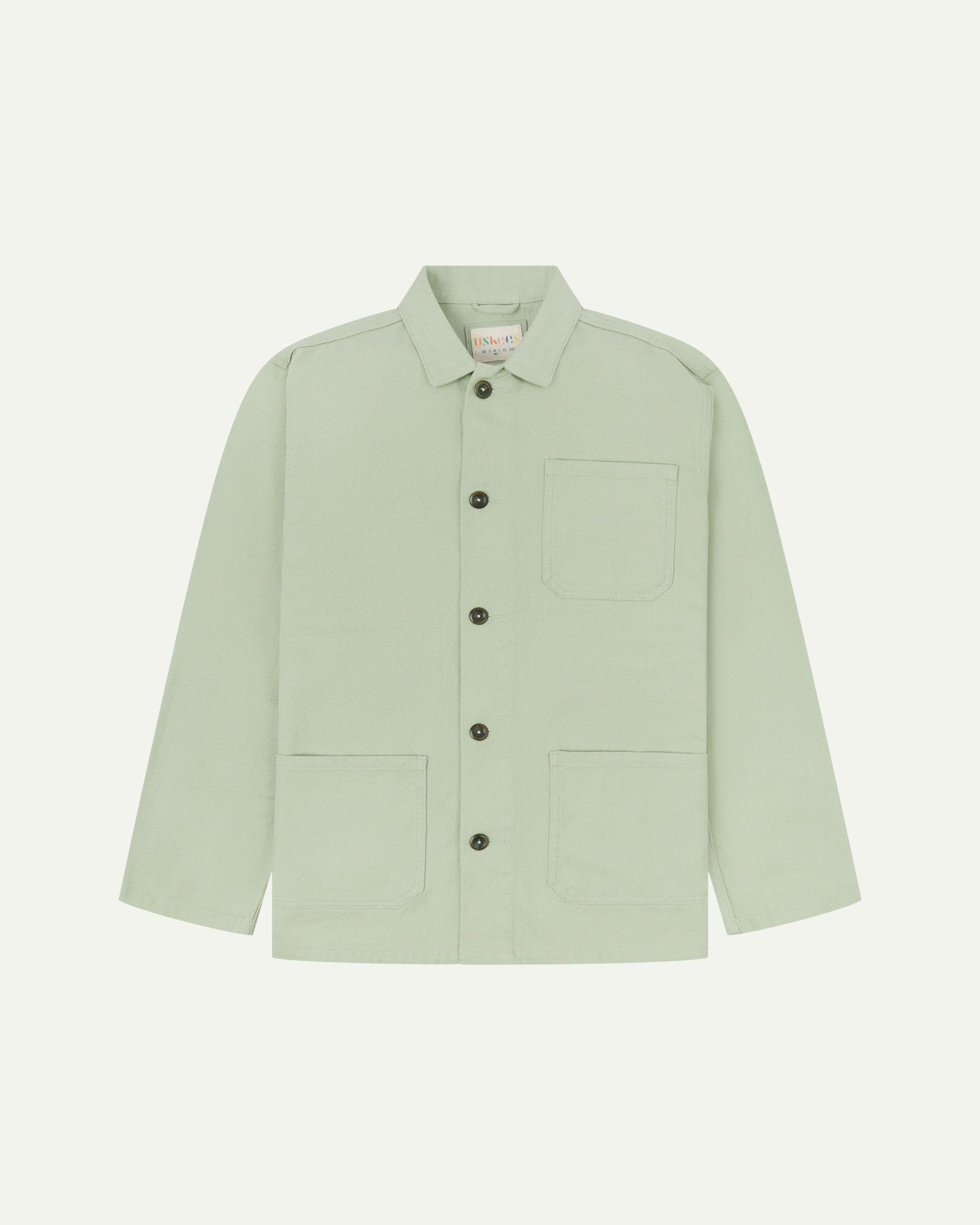 Front flat shot of silver-green, buttoned drill overshirt. Clear view of corozo buttons, 3 pockets and Uskees branding label.