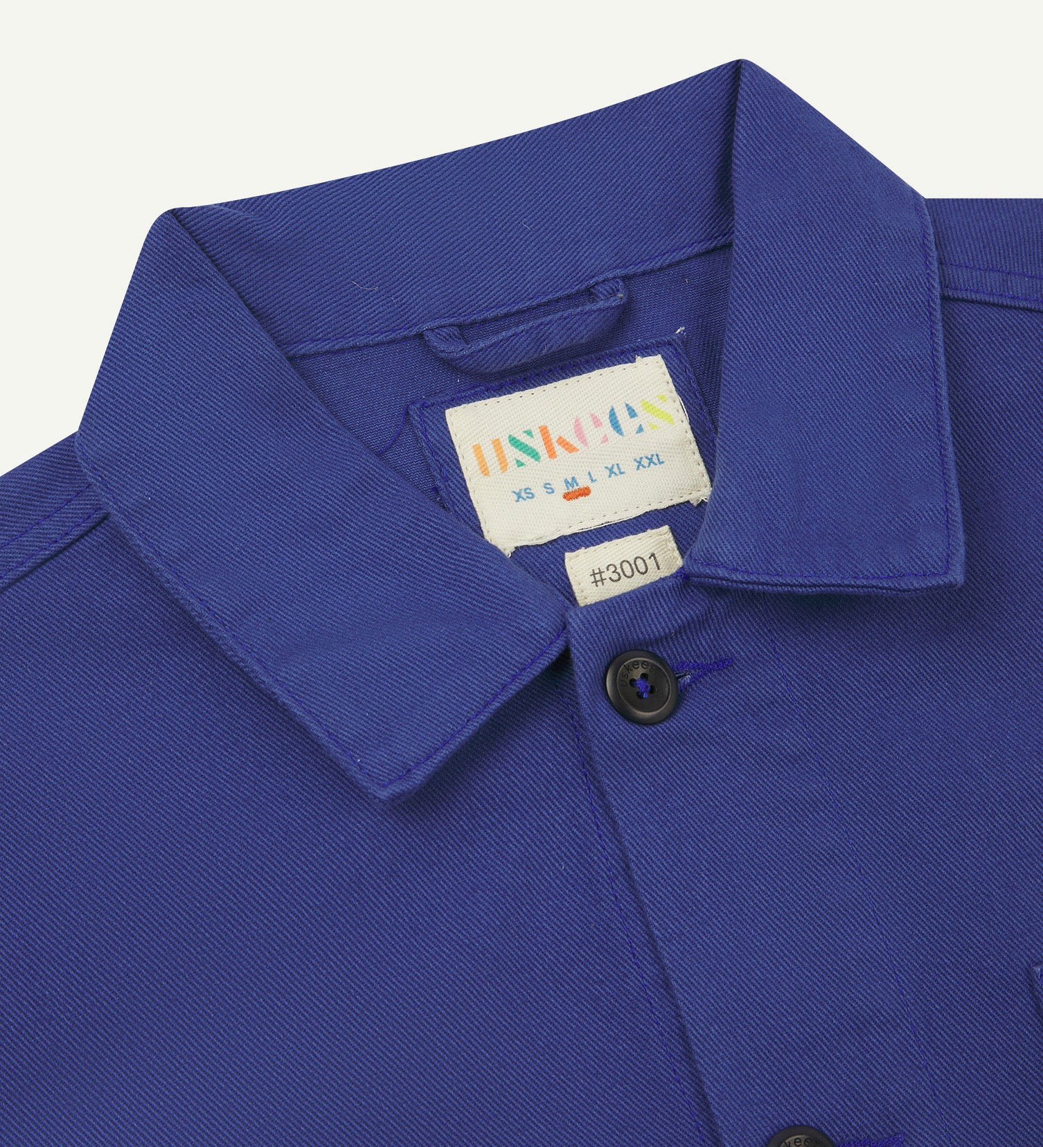 Close-up top-half view of #3001, ultra blue organic cotton drill overshirt. With focus on collar, Uskees brand label and corozo buttons.