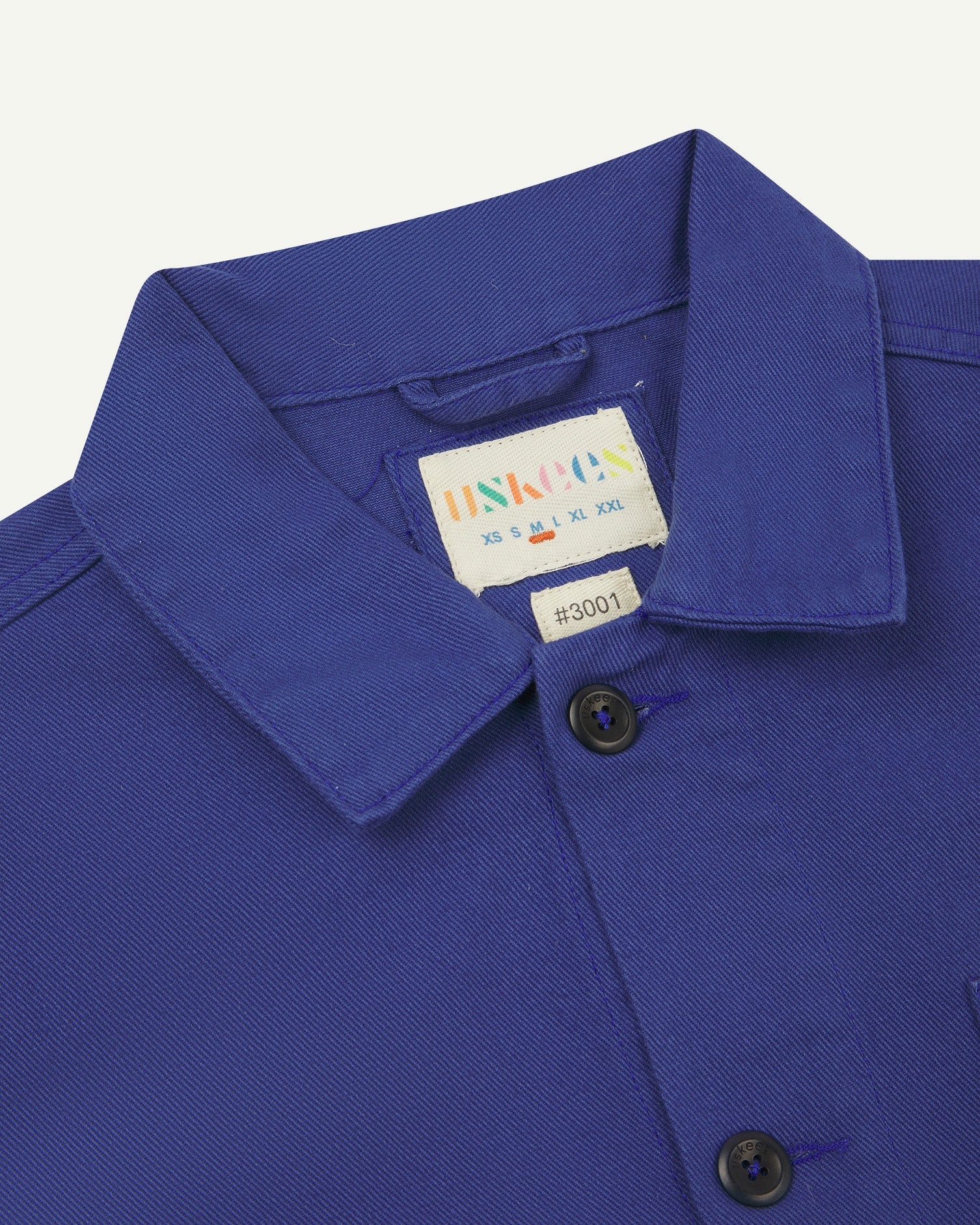 Close-up top-half view of #3001, ultra blue organic cotton drill overshirt. With focus on collar, Uskees brand label and corozo buttons.