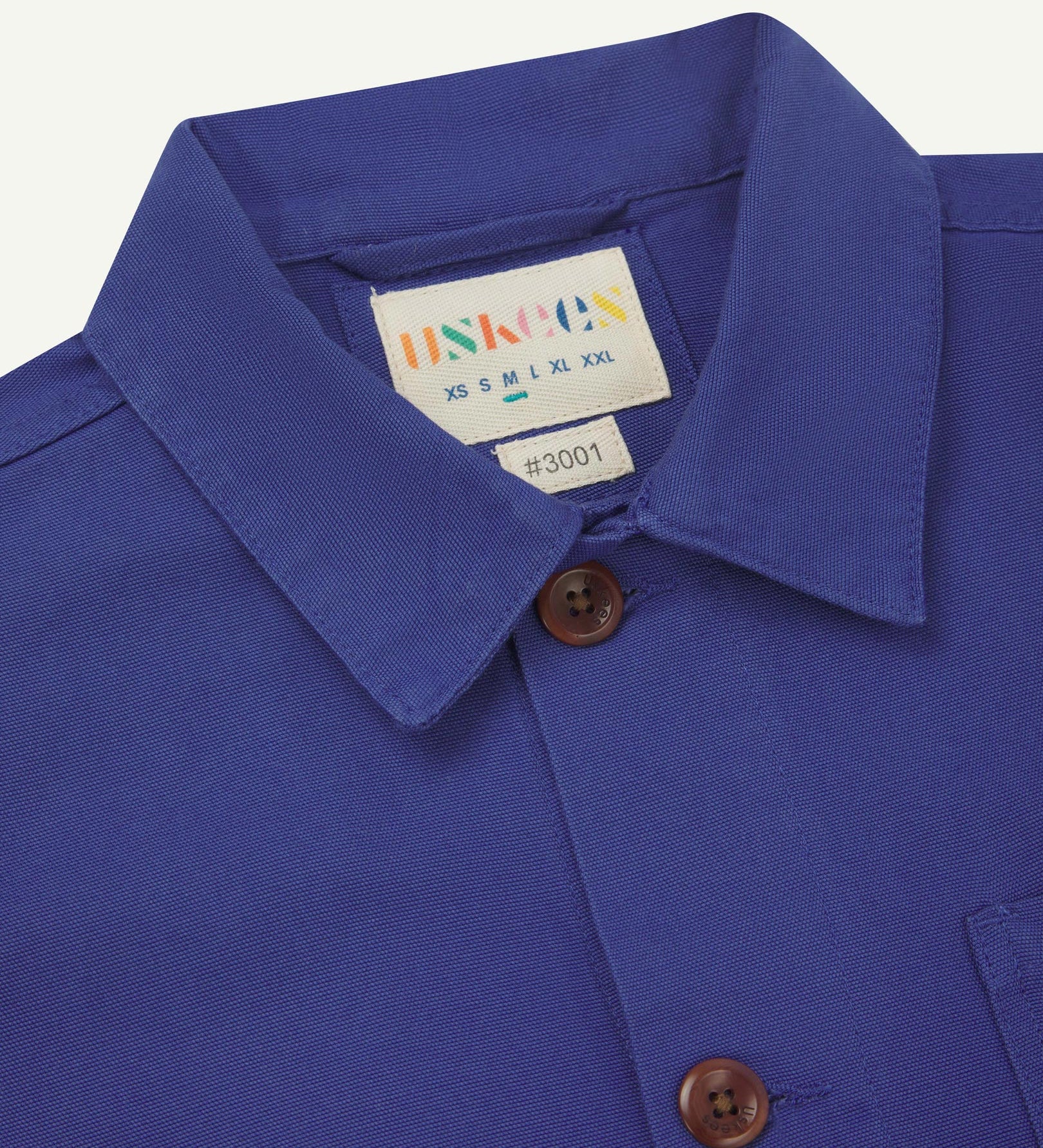 Close-up view of 3001 ultra blue, buttoned organic cotton overshirt from Uskees showing corozo buttons, brand label, collar and hanging hoop.