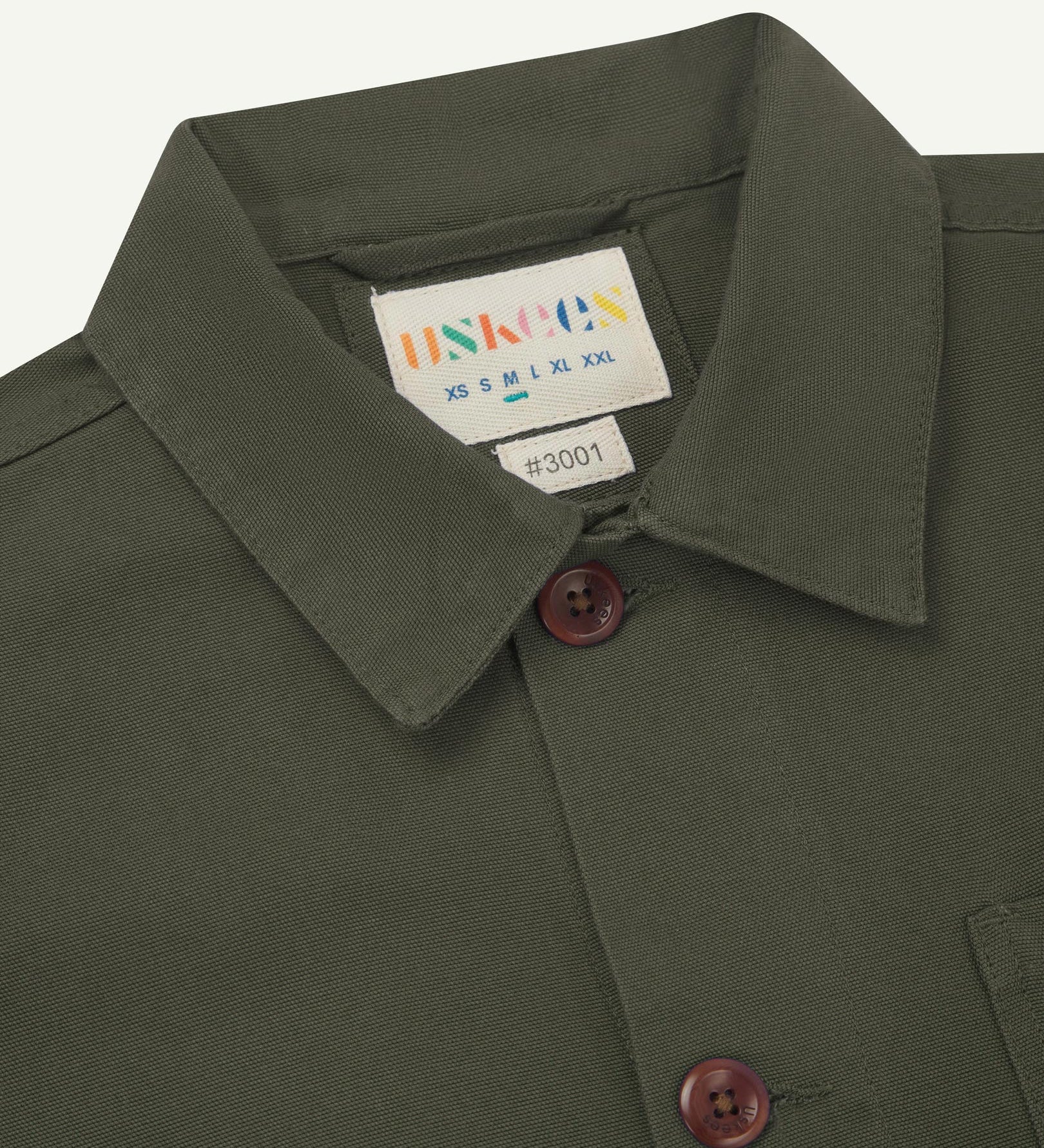 Close-up view of 3001 vine green, buttoned organic cotton overshirt from Uskees showing corozo buttons, brand label, collar and hanging hoop.
