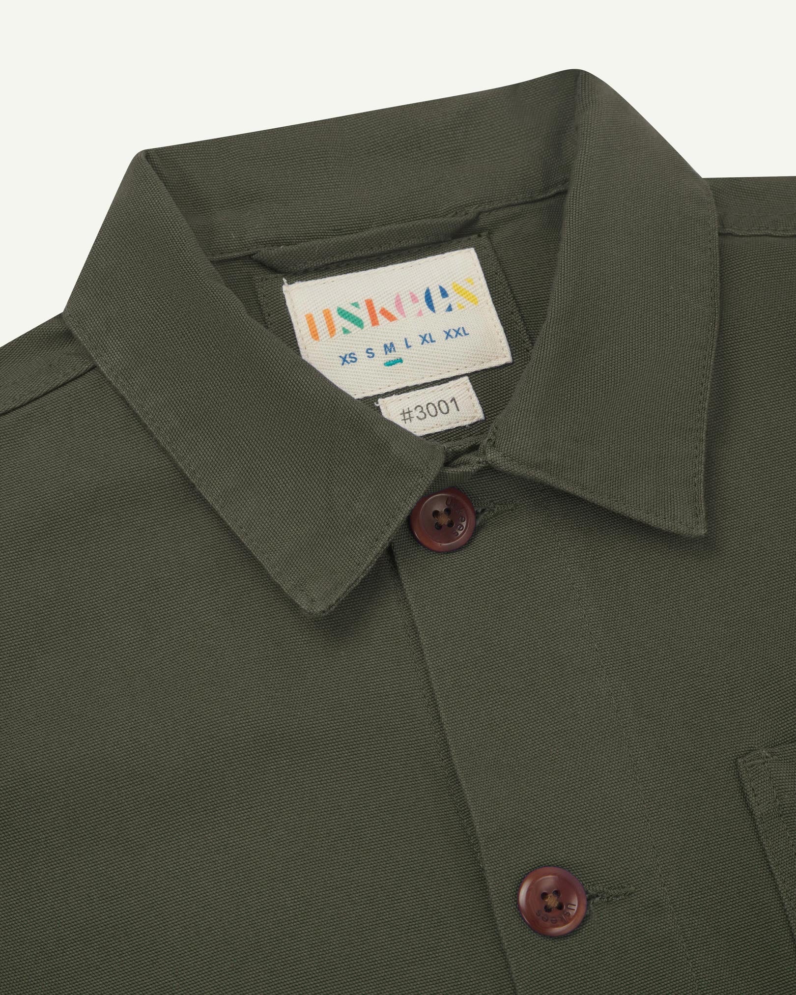 Close-up view of 3001 vine green, buttoned organic cotton overshirt from Uskees showing corozo buttons, brand label, collar and hanging hoop.