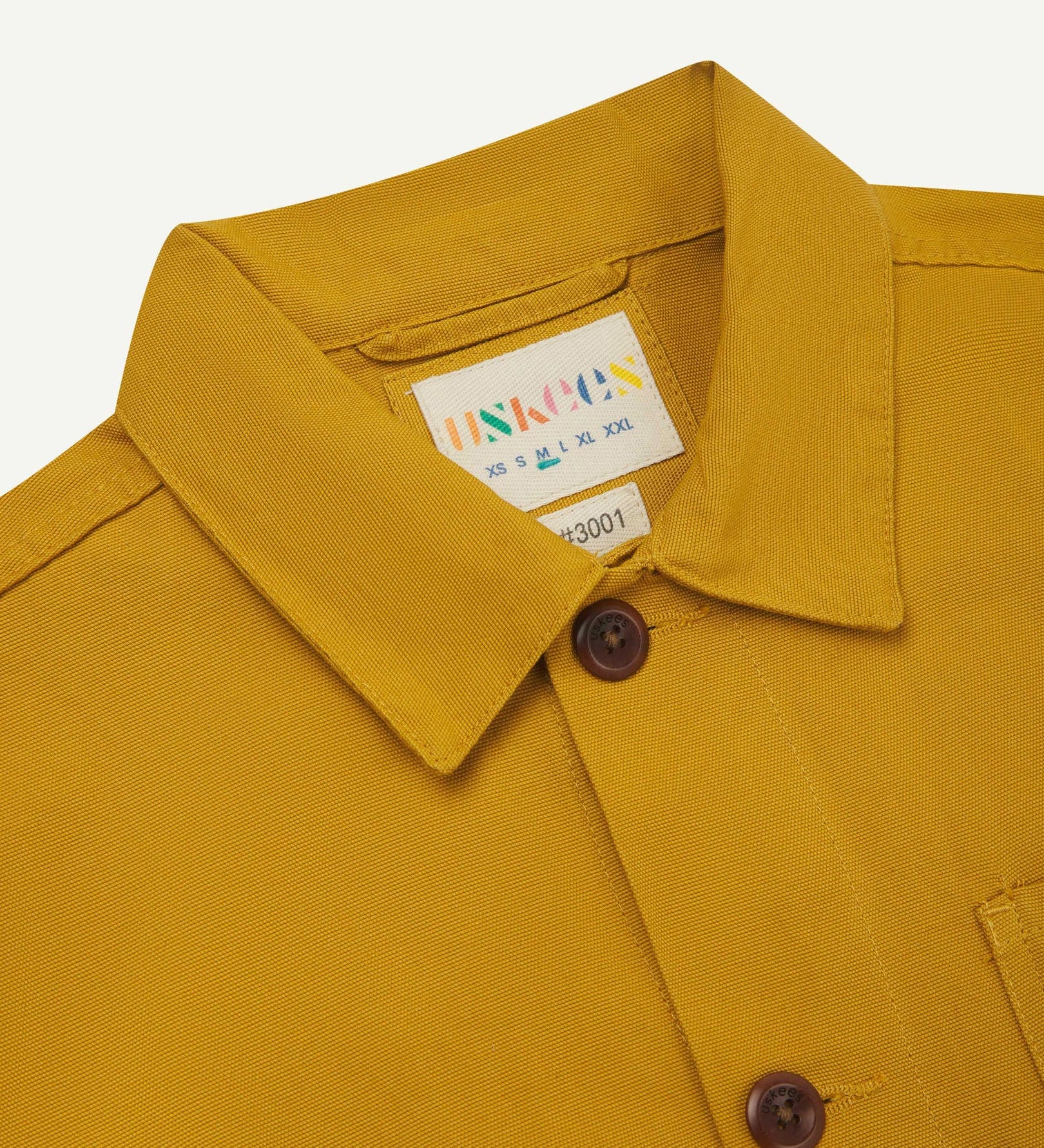 Close-up view of 3001 yellow, buttoned organic cotton overshirt from Uskees showing corozo buttons, brand label, collar and hanging hoop.