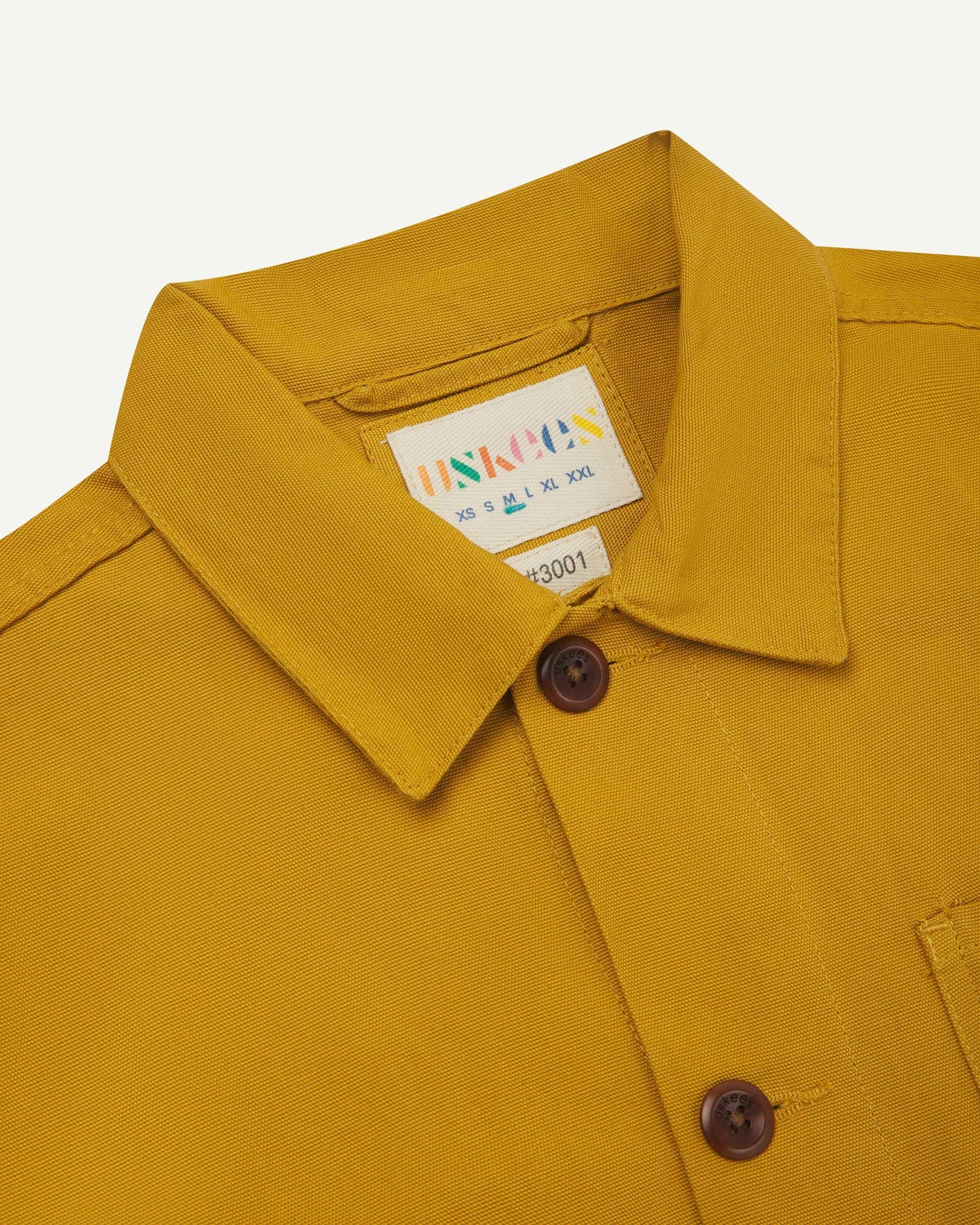 Close-up view of 3001 yellow, buttoned organic cotton overshirt from Uskees showing corozo buttons, brand label, collar and hanging hoop.