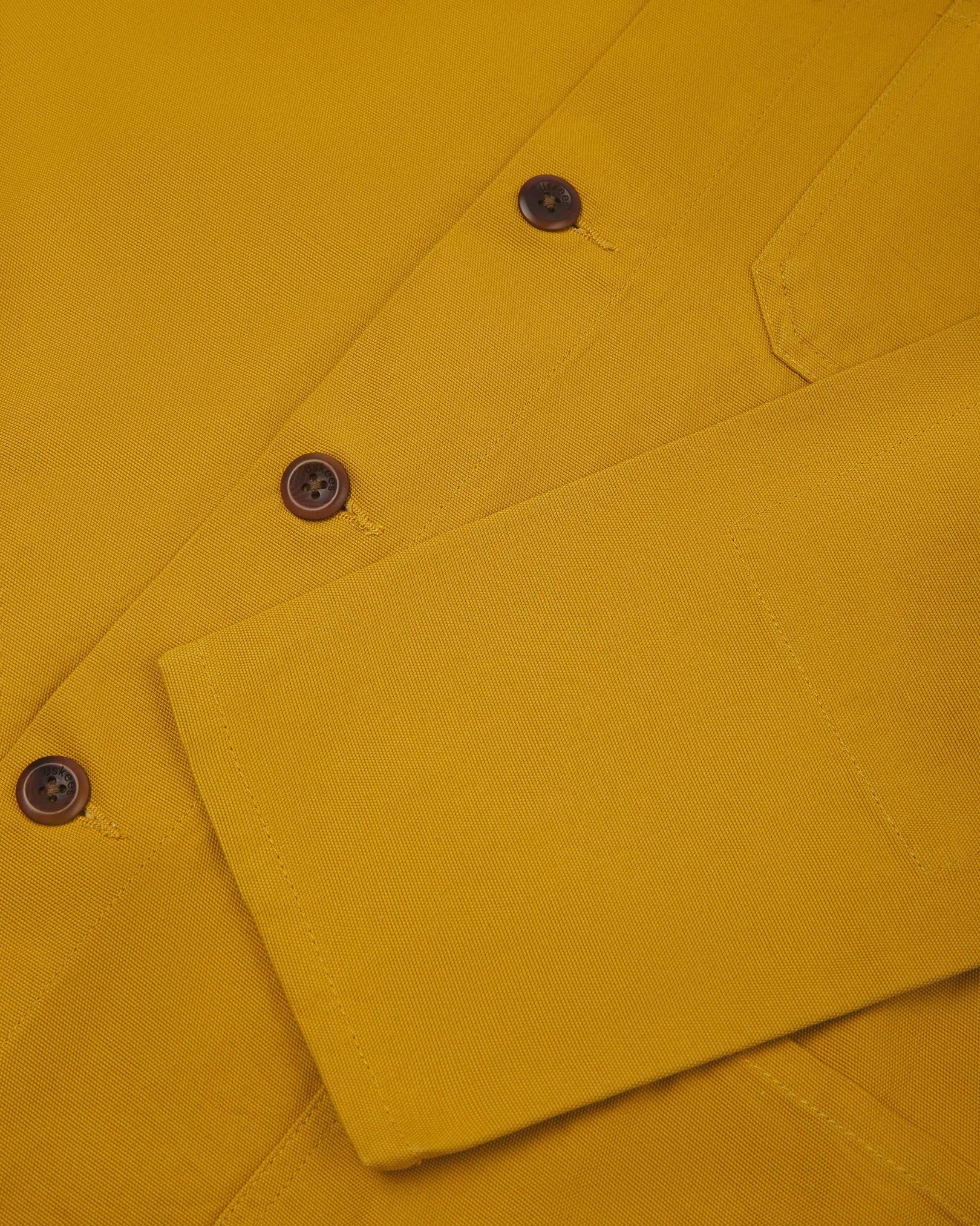 Closer view of mid section of yellow, buttoned organic cotton overshirt from Uskees. Focus on cuff, pockets and corozo buttons.