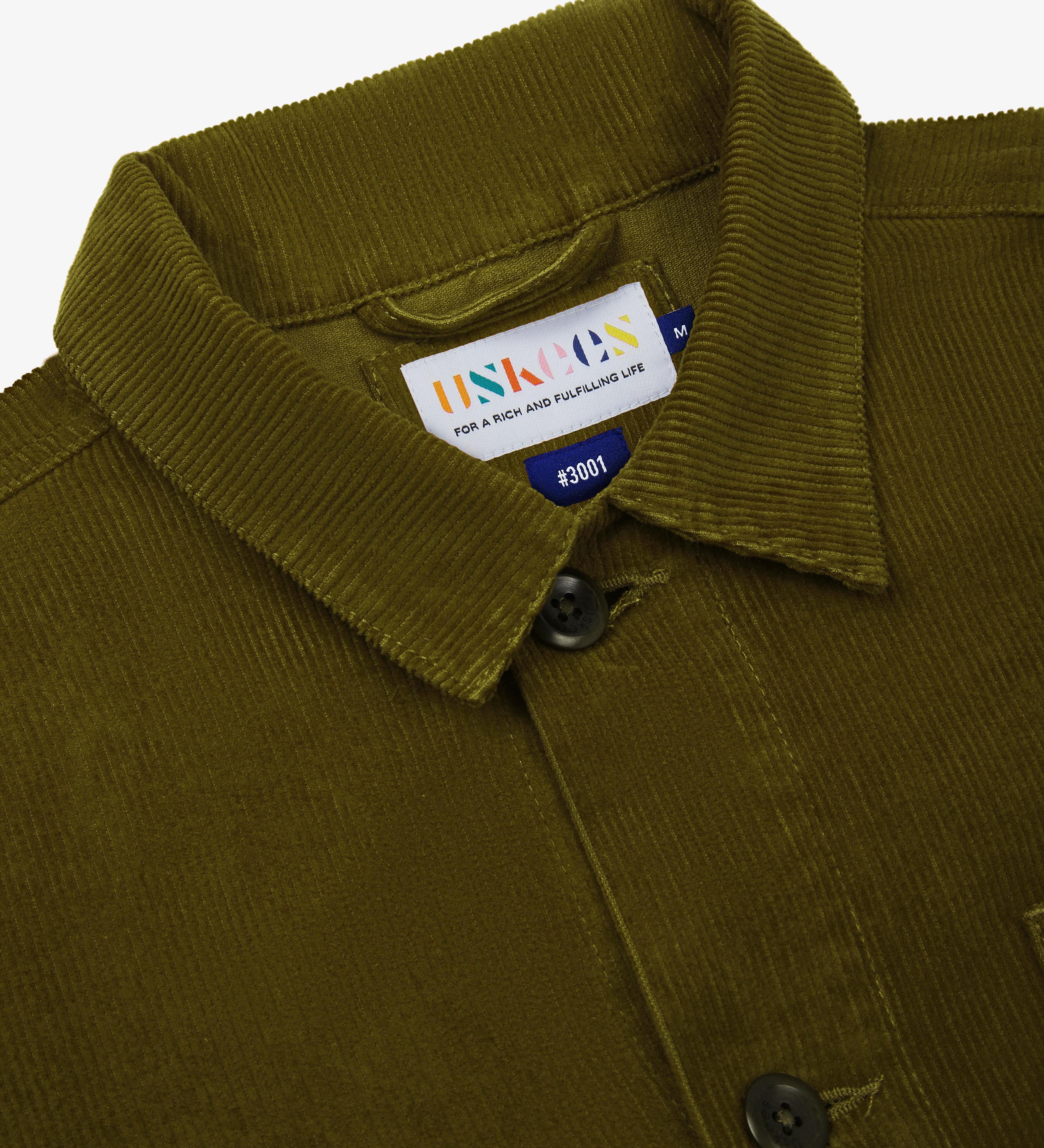 Close-up of a green corduroy overshirt showing an Uskees brand label on a white background