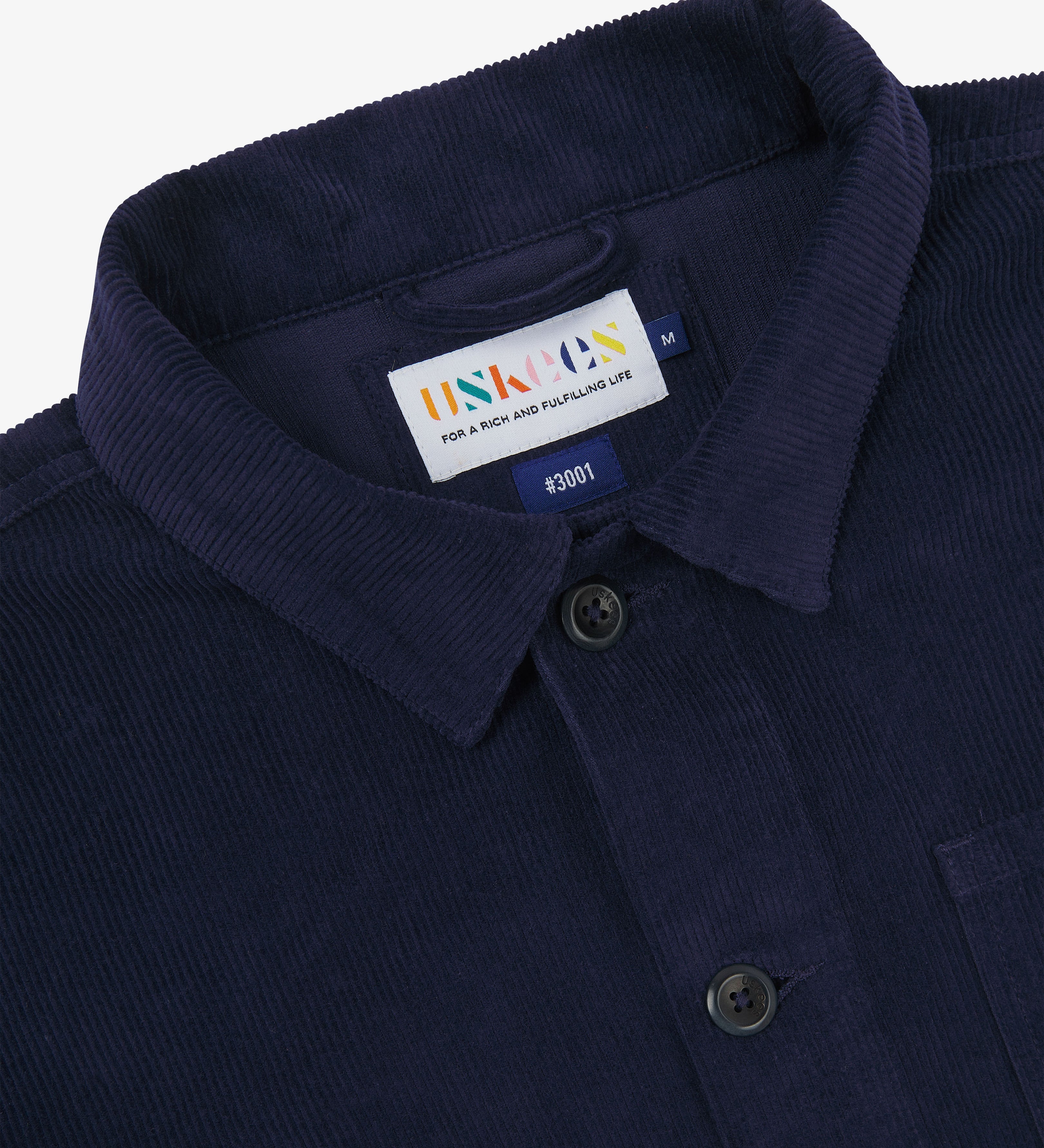 close-up of indigo blue cord overshirt collar with a visible Uskees brand label on a white background