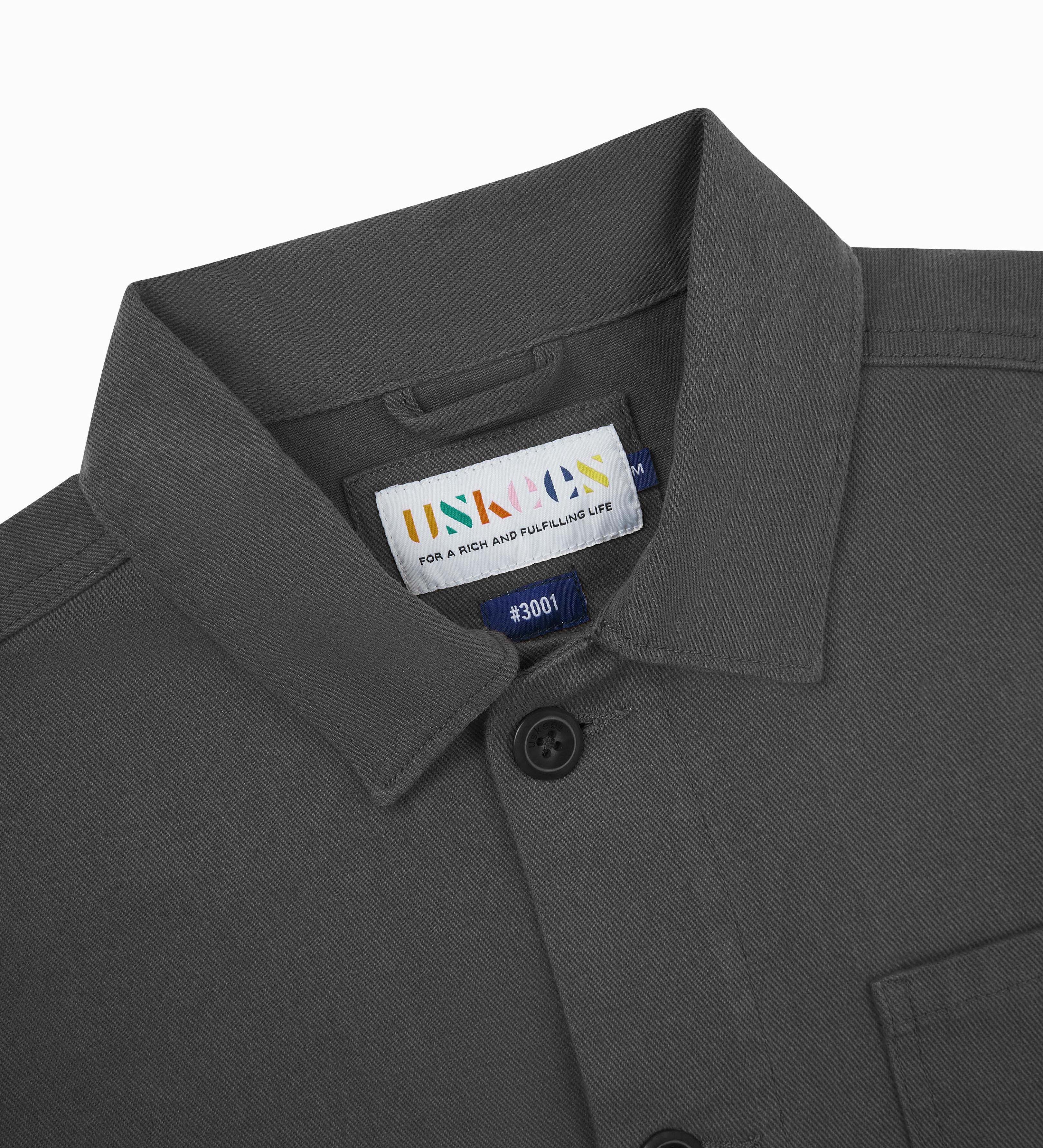 Close-up top-half view of #3001, charcoal-grey organic cotton drill overshirt. With focus on collar, Uskees brand label and corozo buttons.