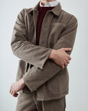 A neck to thigh shot of a male model wearing a taupe-brown corduroy overshirt jacket over a burgundy knit sweater and white collared shirt,  against a light gray studio background.