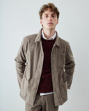 A male model wearing an Uskees light brown corduroy utility jacket over a burgundy knit sweater and white collared shirt, standing against a light gray studio background.