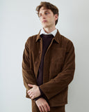 A male model wearing a brown corduroy utility jacket over a dark red ribbed sweater and white collared shirt, standing against a plain light gray studio background.