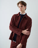 A male model wearing an Uskees dark red wine colour corduroy shirt jacket over a navy blue ribbed sweater and white collared shirt, standing against a light gray studio background.
