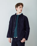 A male model wearing a navy blue corduroy utility shirt jacket over a green/blue patterned sweater and white collared shirt, standing against a light gray studio background.