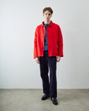 A man stands in a studio wearing a bright red button-front jacket over a blue knit top, dark navy trousers, and black shoes against a plain light gray wall and concrete floor.