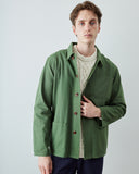 A man wearing an Uskees mid-green utility jacket over a cream cable-knit sweater and navy trousers, standing against a light gray studio background.