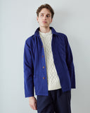 A male model wearing a deep indigo utility jacket over a cream cable-knit sweater and navy trousers, standing against a light gray studio background.