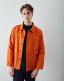 A male model wearing a bright orange Uskees utility jacket over a dark navy sweater and navy trousers, standing against a plain light gray studio background.