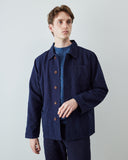 A male model wearing an Uskees navy blue utility jacket over a textured blue sweater and dark trousers, standing against a light gray studio background.