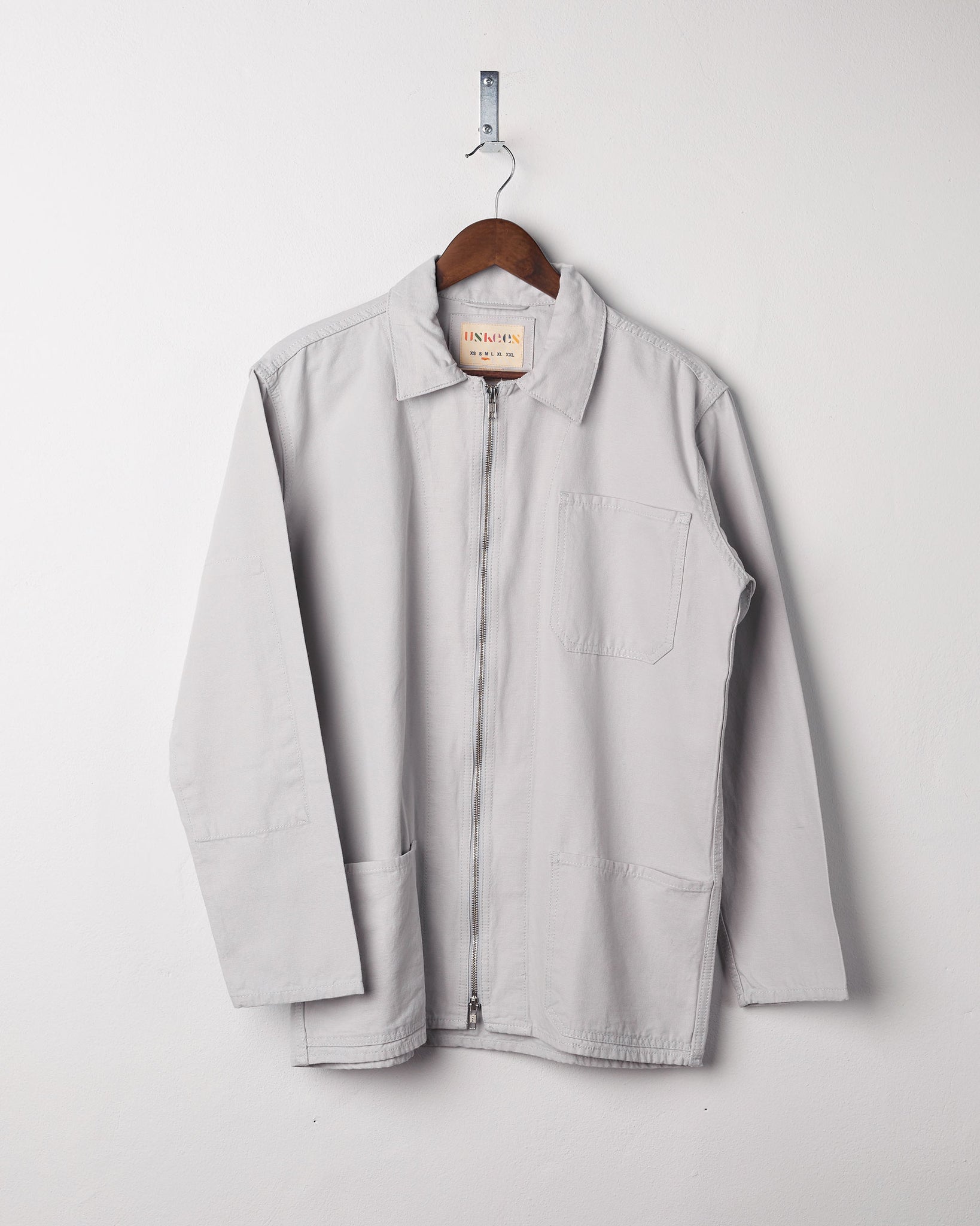 Front view of light-grey, organic cotton zip front jacket from Uskees presented on hanger with neutral backdrop.
