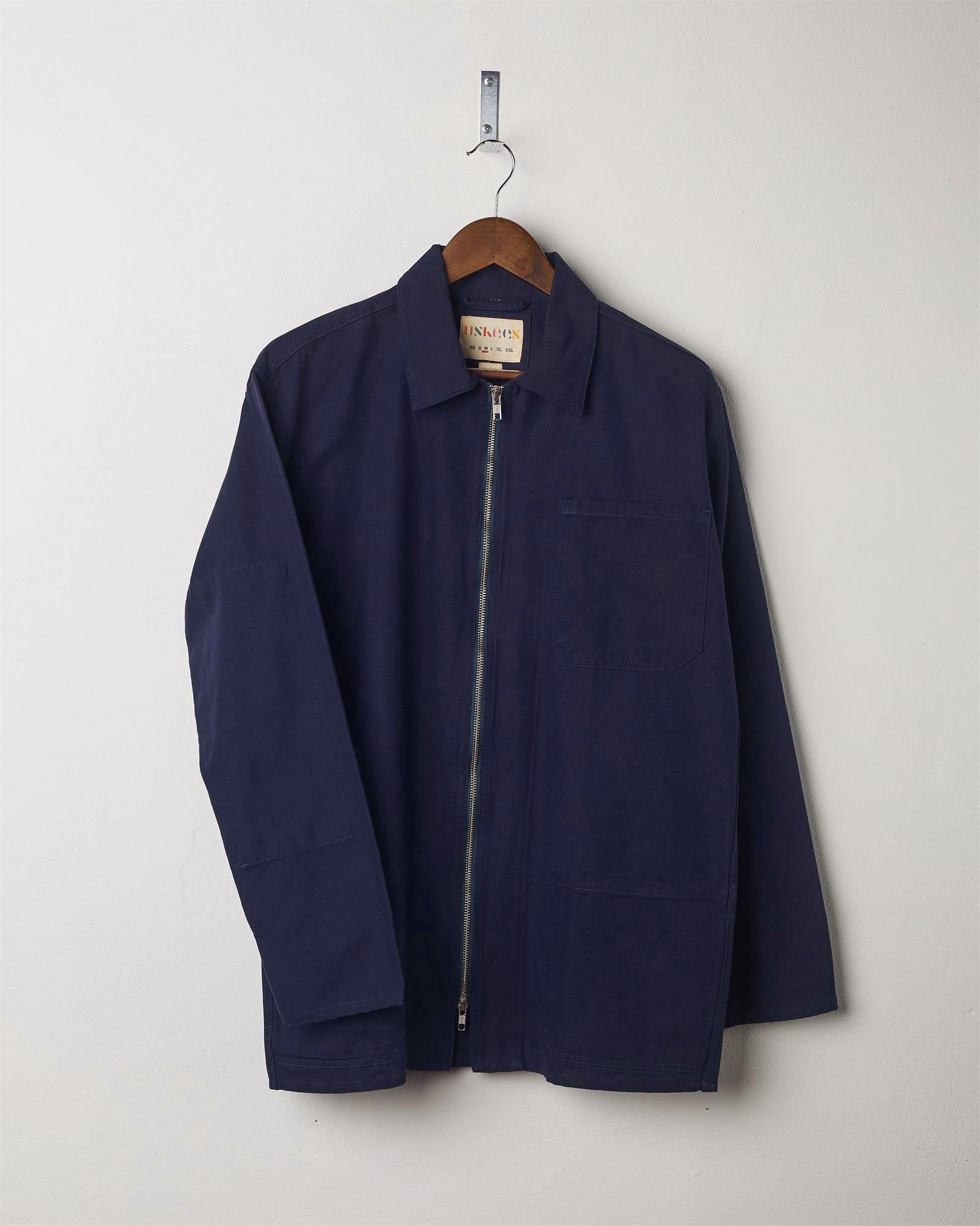 Front view of midnight blue, organic cotton zip front jacket from Uskees presented on hanger with neutral backdrop.