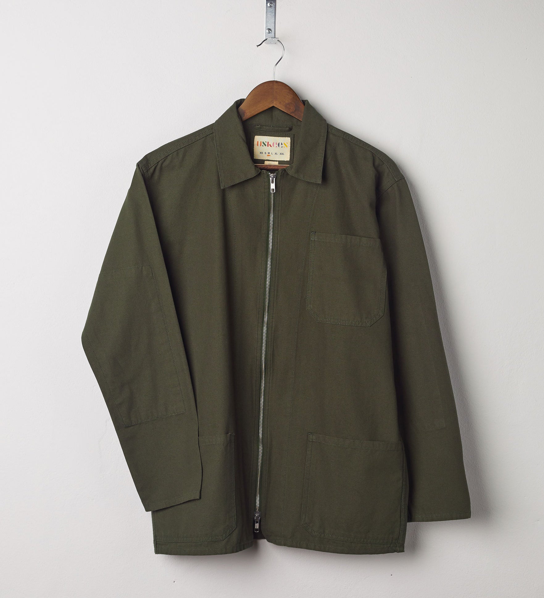 Front view of vine green, organic cotton zip front jacket from Uskees presented on hanger with neutral backdrop.