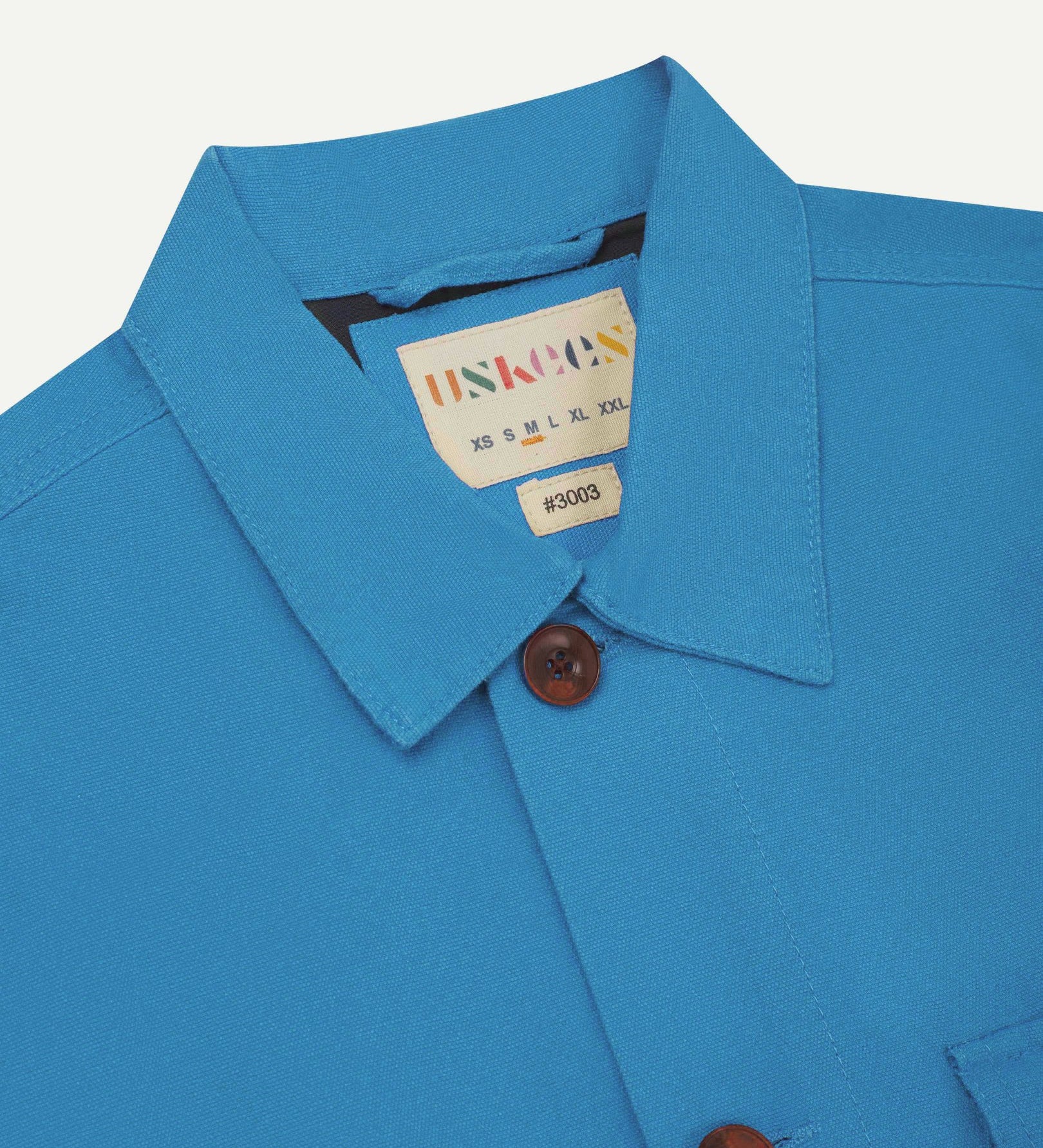 Close-up top-half view of #3003, bright blue organic cotton workshirt. With focus on collar, Uskees brand label and corozo buttons.