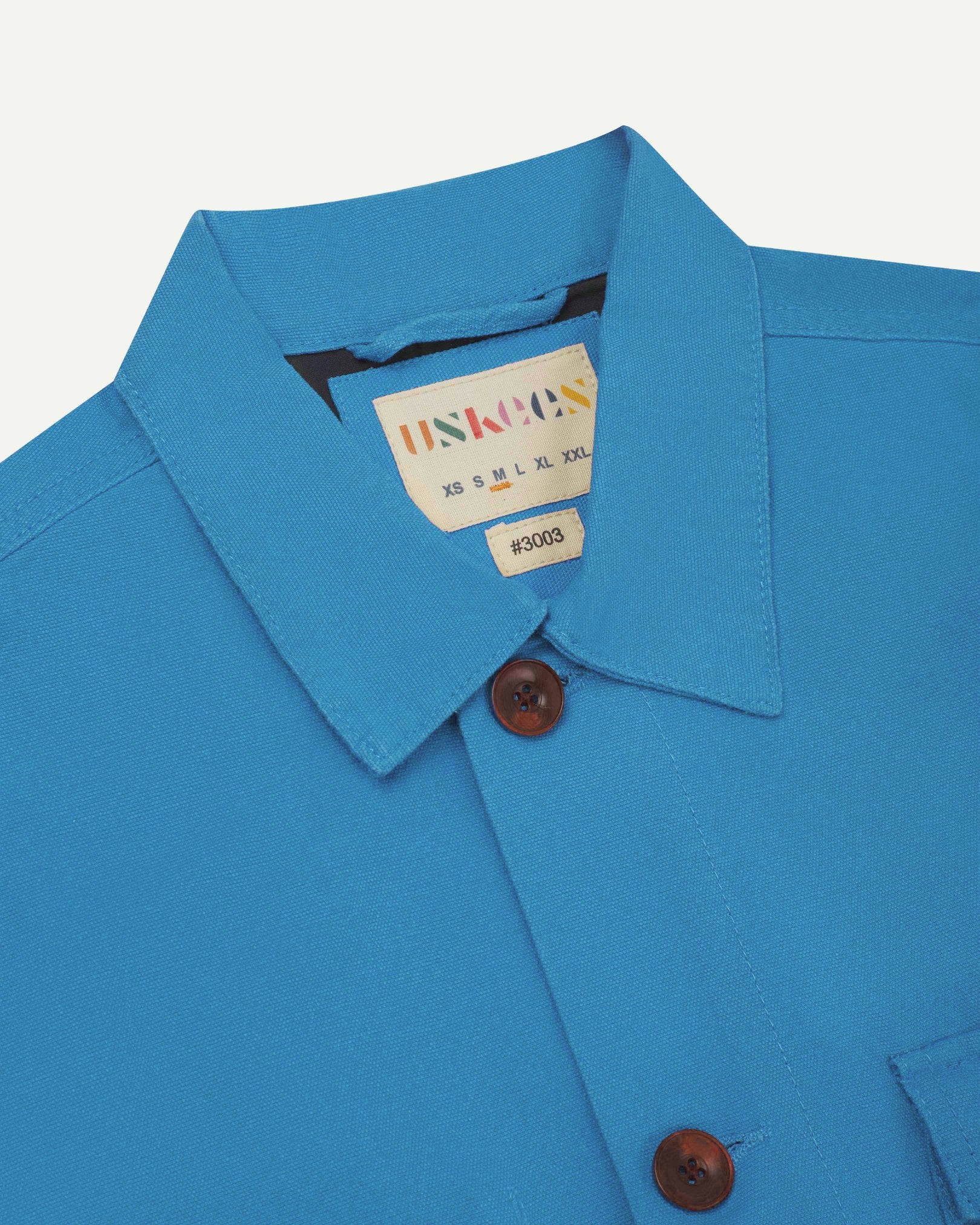 Close-up top-half view of #3003, bright blue organic cotton workshirt. With focus on collar, Uskees brand label and corozo buttons.