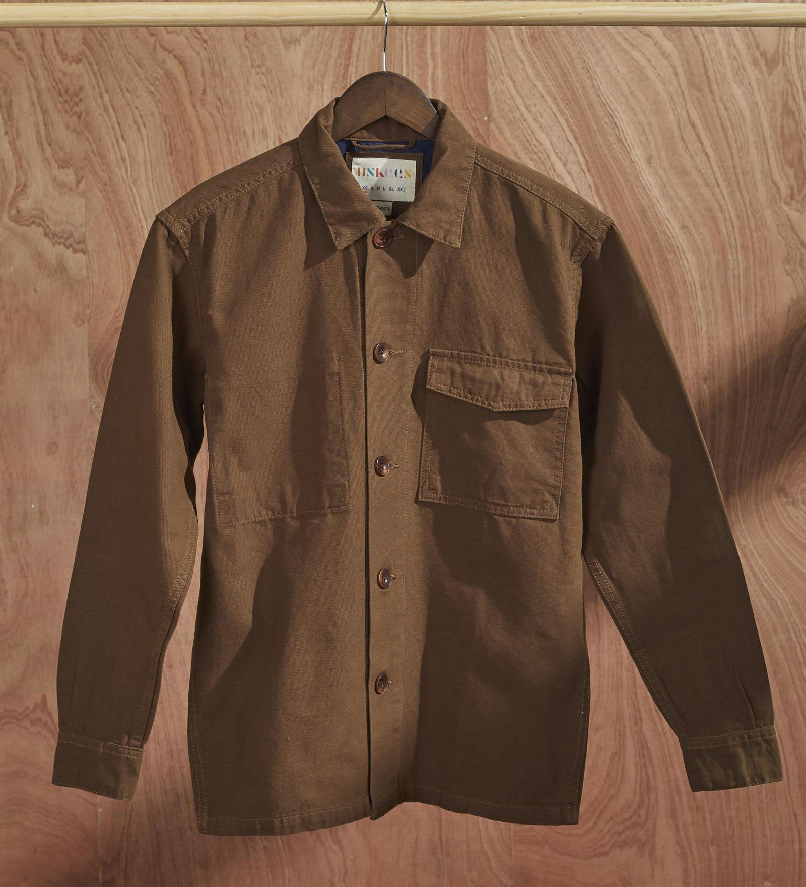 Front view of brown coloured, buttoned workshirt from Uskees presented on hanger with wooden backdrop.