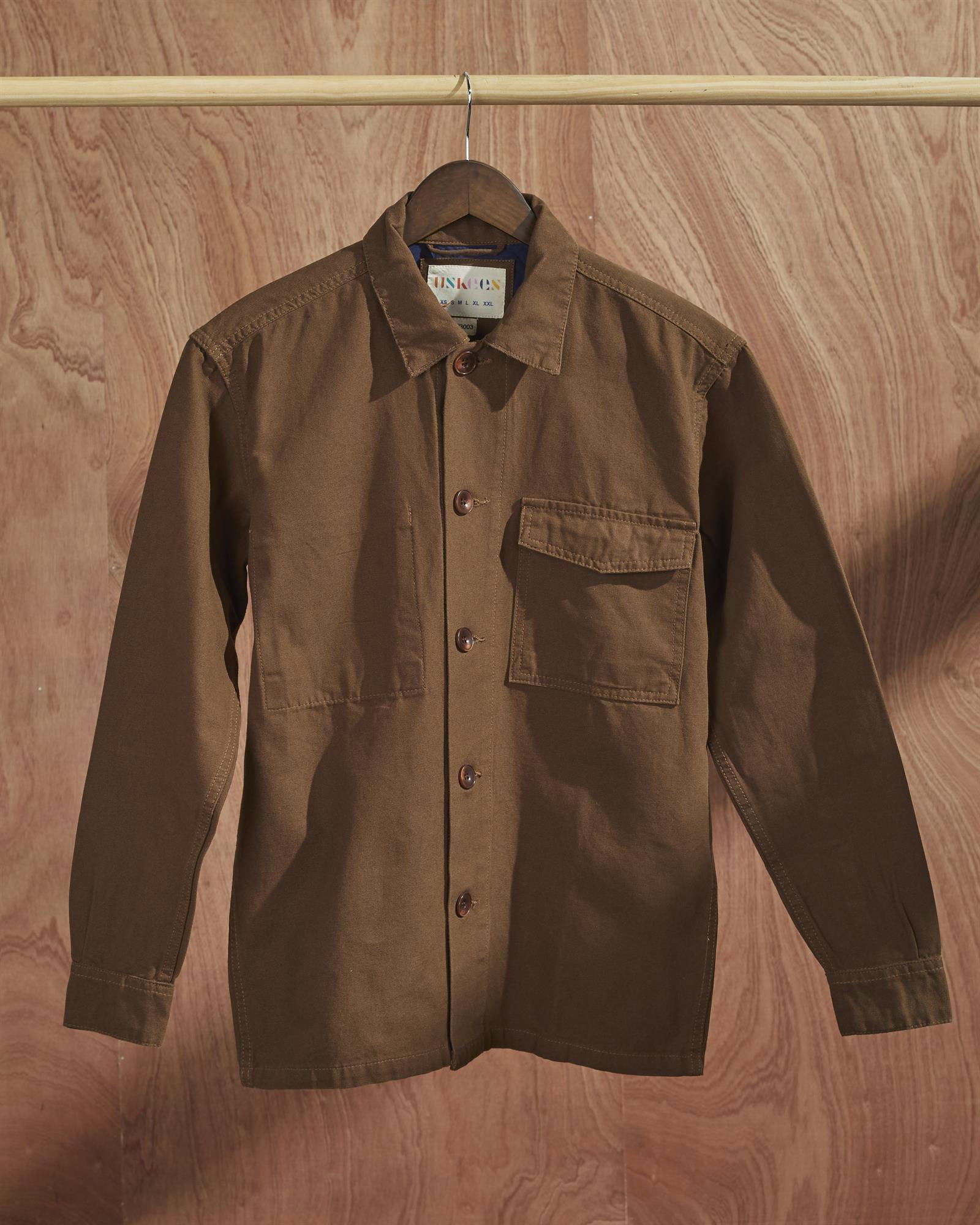 Front view of brown coloured, buttoned workshirt from Uskees presented on hanger with wooden backdrop.