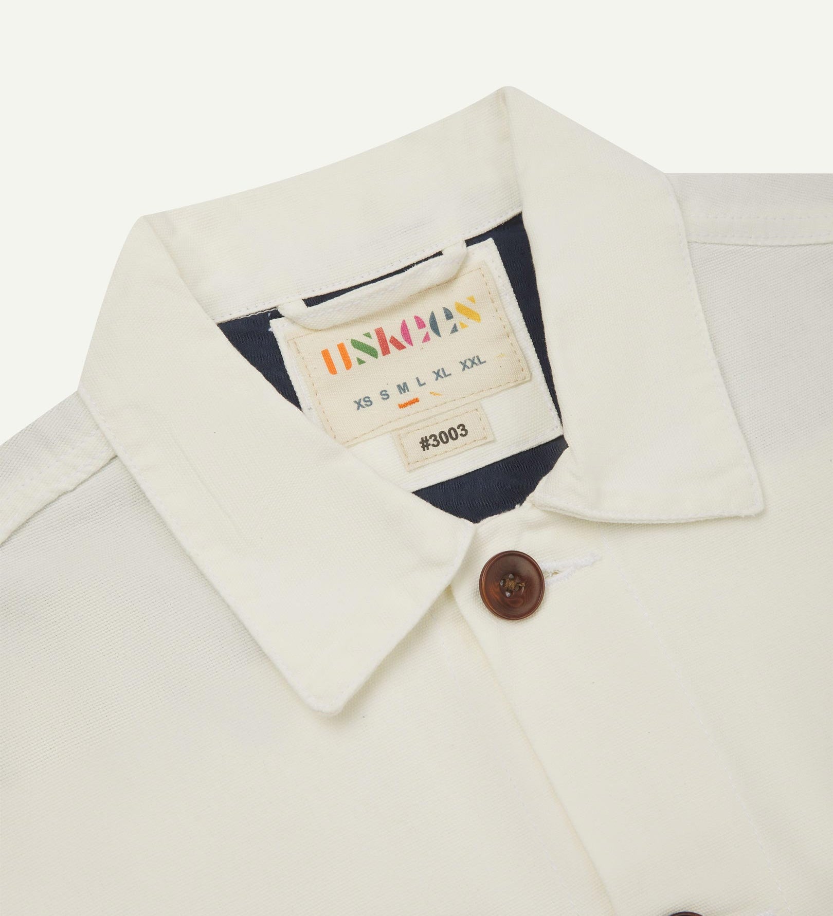 Close-up top-half view of #3003, cream organic cotton workshirt. With focus on collar, Uskees brand label and corozo buttons.