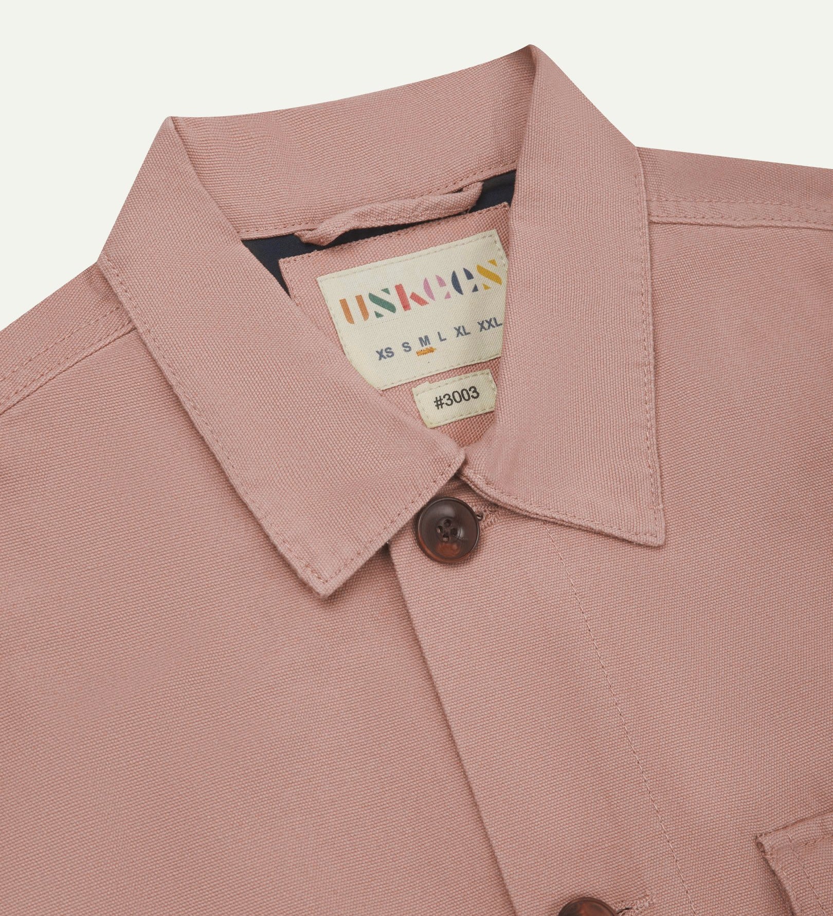 Close-up top-half view of #3003, dusty pink organic cotton workshirt. With focus on collar, contrasting yoke, Uskees brand label and corozo buttons.