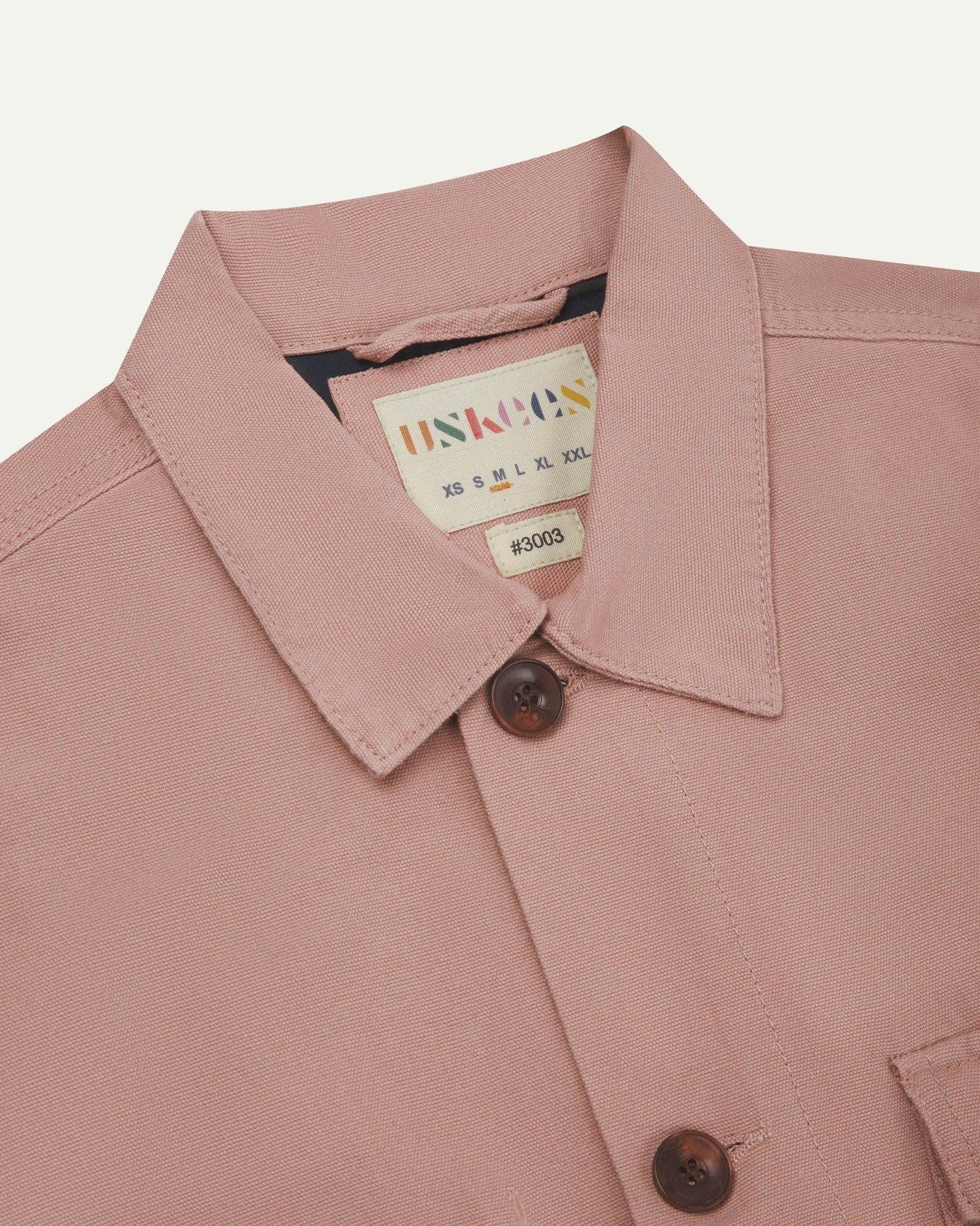 Close-up top-half view of #3003, dusty pink organic cotton workshirt. With focus on collar, contrasting yoke, Uskees brand label and corozo buttons.