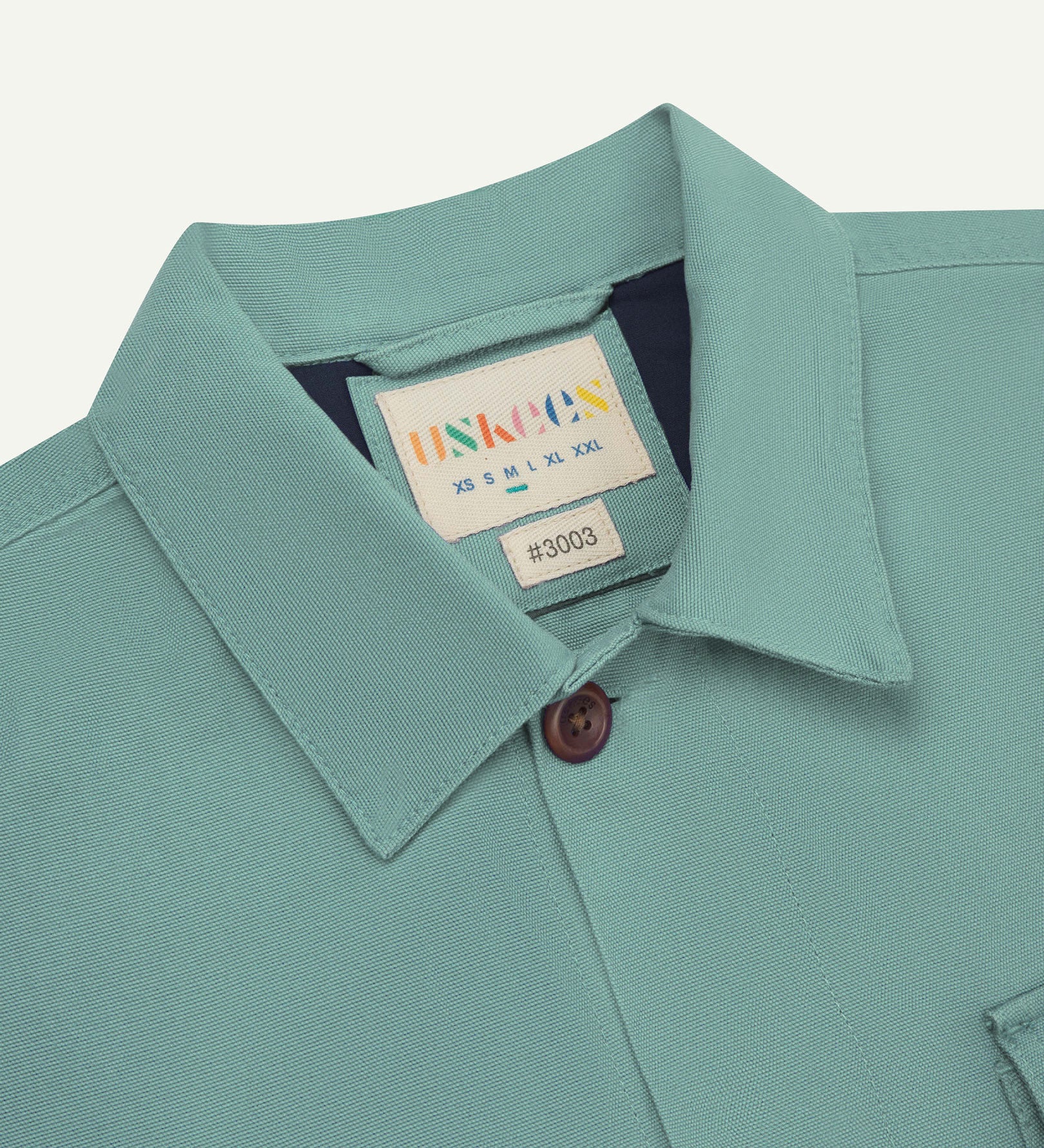 Close-up top-half view of #3003, eucalyptus (mint-green) organic cotton workshirt. With focus on collar, Uskees brand label and corozo buttons.