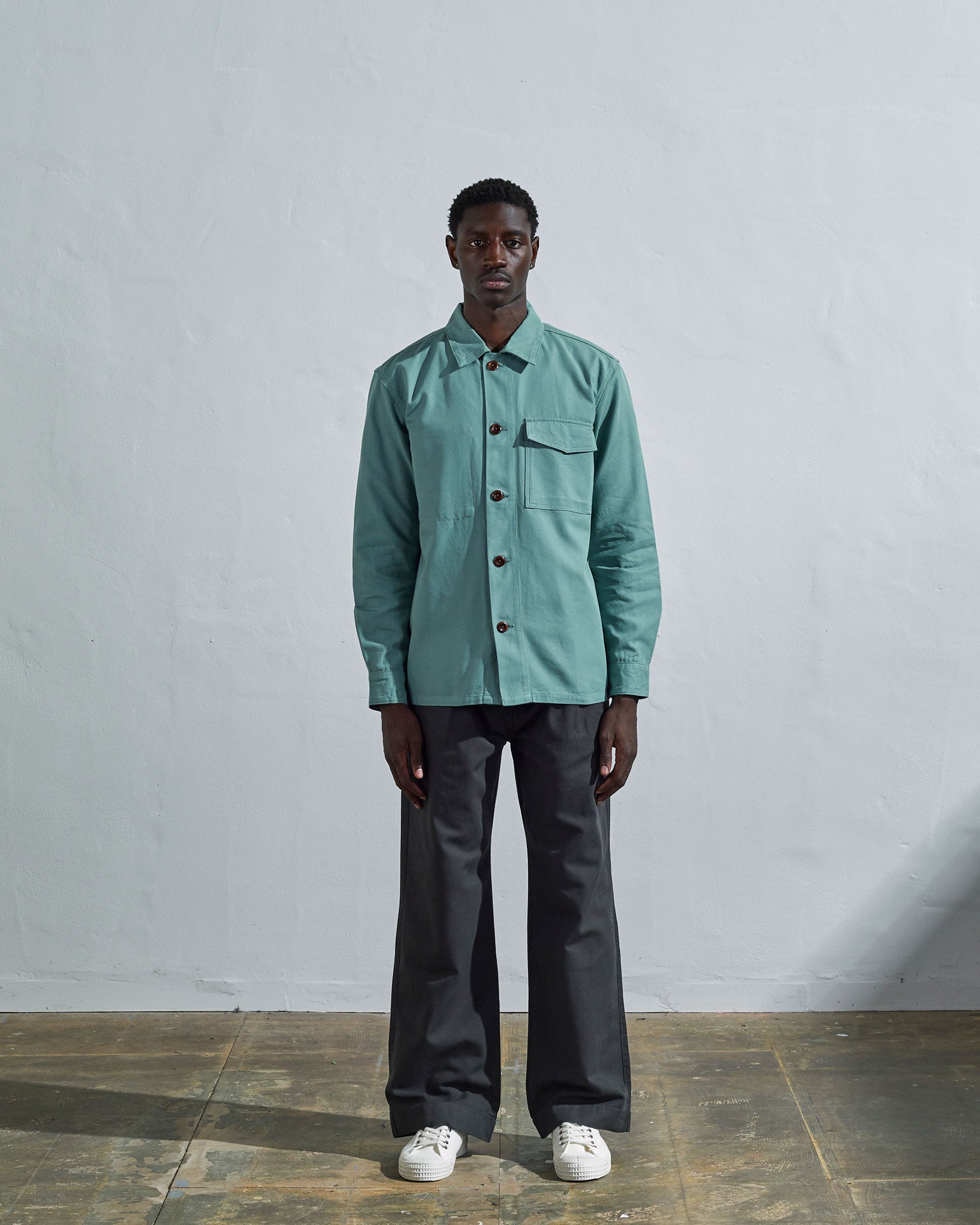 Full-length view of model wearing Uskees #3003 green-blue 'eucalyptus' workshirt illustrating utilitarian silhouette. Paired with dark Uskees pants.