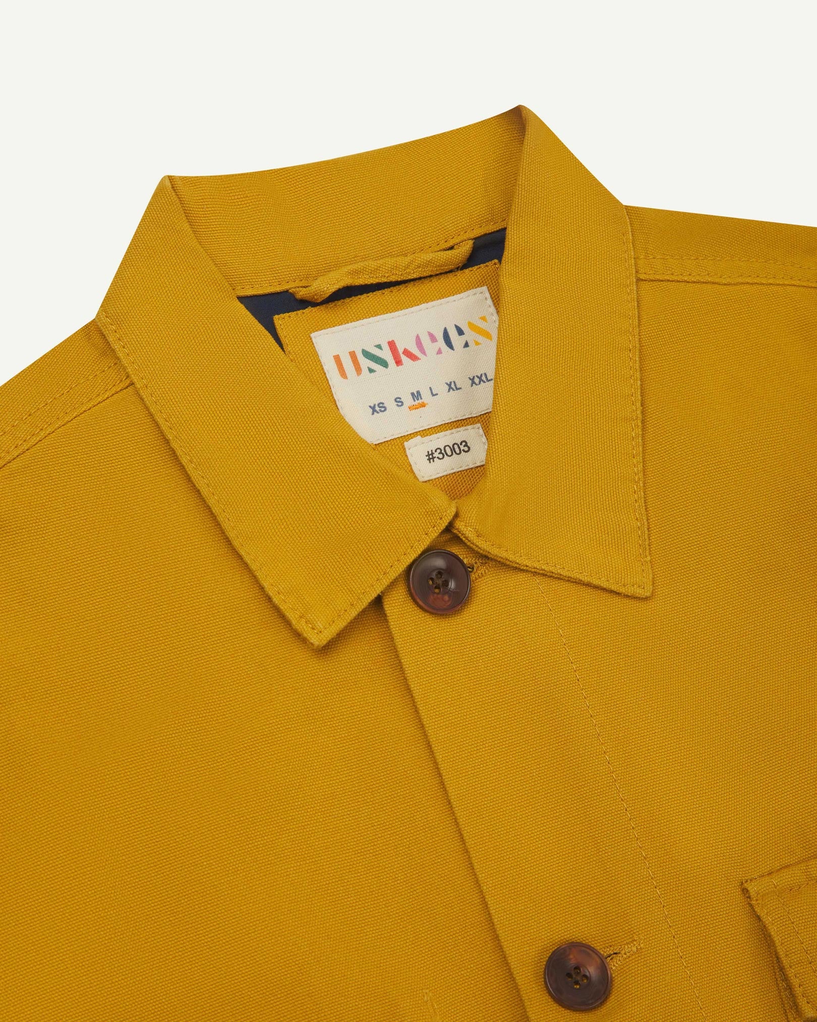 Close-up top-half view of #3003, yellow organic cotton workshirt. With focus on collar, Uskees brand label and corozo buttons.
