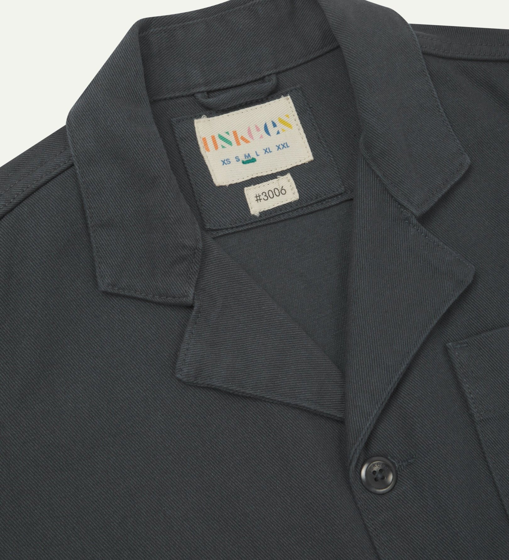 Close-up top-half view of #3006, charcoal-grey organic cotton-drill blazer. With focus on collar, lapels, Uskees brand label and corozo buttons.