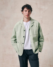 Male model wearing silver-green unbuttoned organic drill blazer from Uskees paired with a white Tee and blue trousers. 