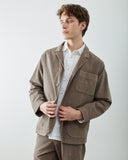 A male model wearing an Uskees taupe-brown corduroy jacket over a white button-down shirt, paired with matching trousers, photographed against a light gray studio background.
