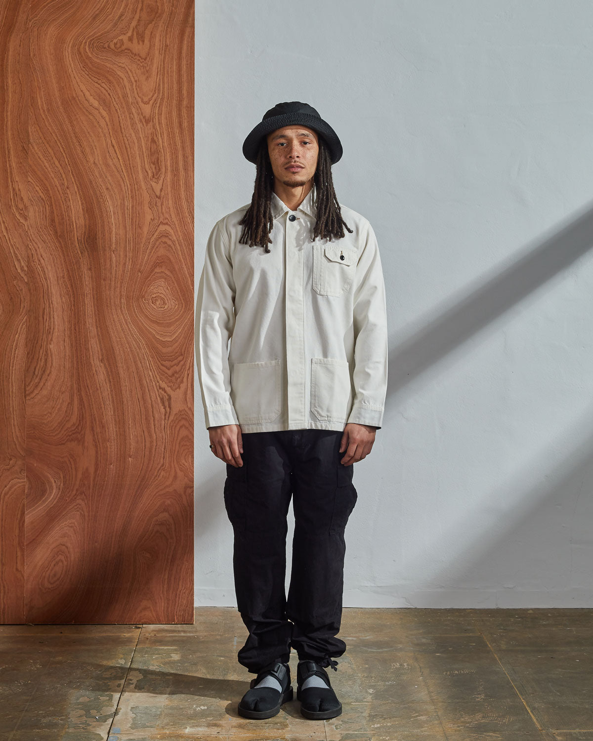 Full-length front model shot of Uskees #3011 cream Men's Overshirt with hidden front buttons. Paired with dark cargo pants and matching hat.