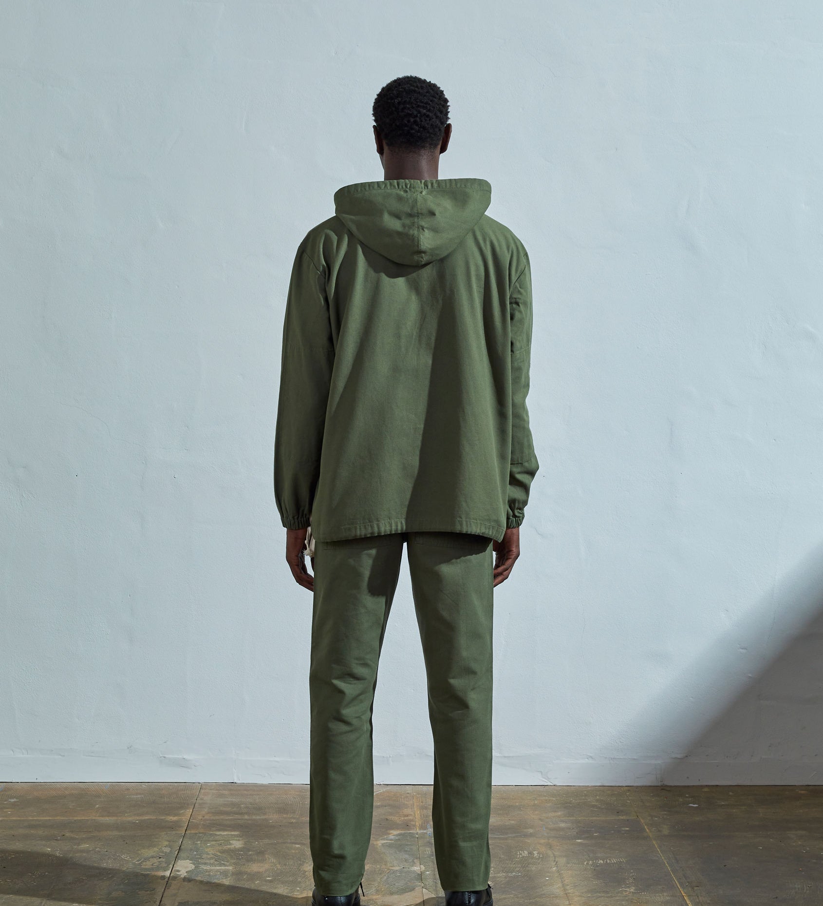 Full-length back view wearing Uskees 'coriander-green' coloured buttoned smock, with view of hood, reinforced elbows and boxy silhouette.