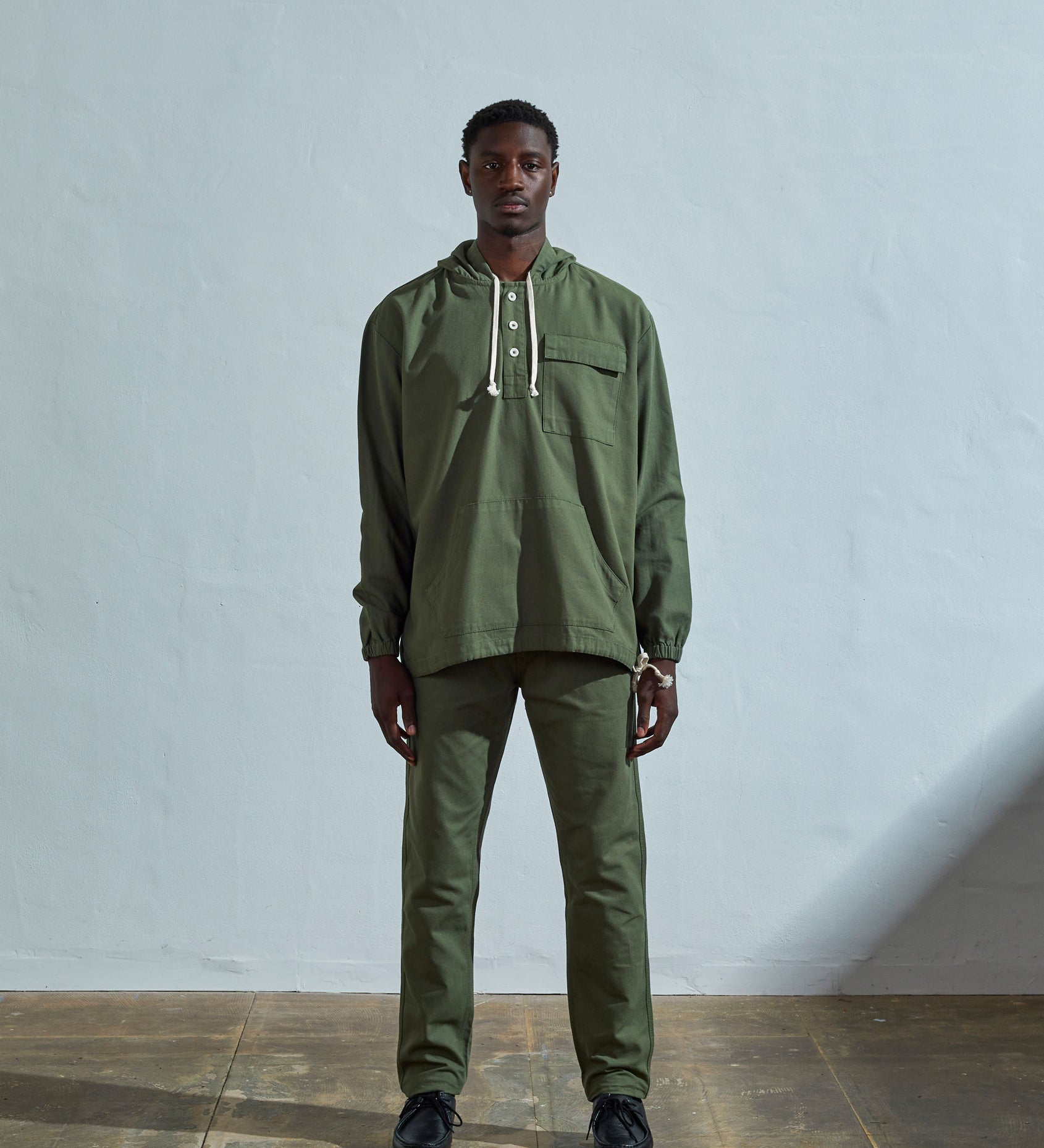 Full-length front view wearing Uskees 'coriander-green' smock, with view of corozo buttons, ribbed cuffs and drawstrings.