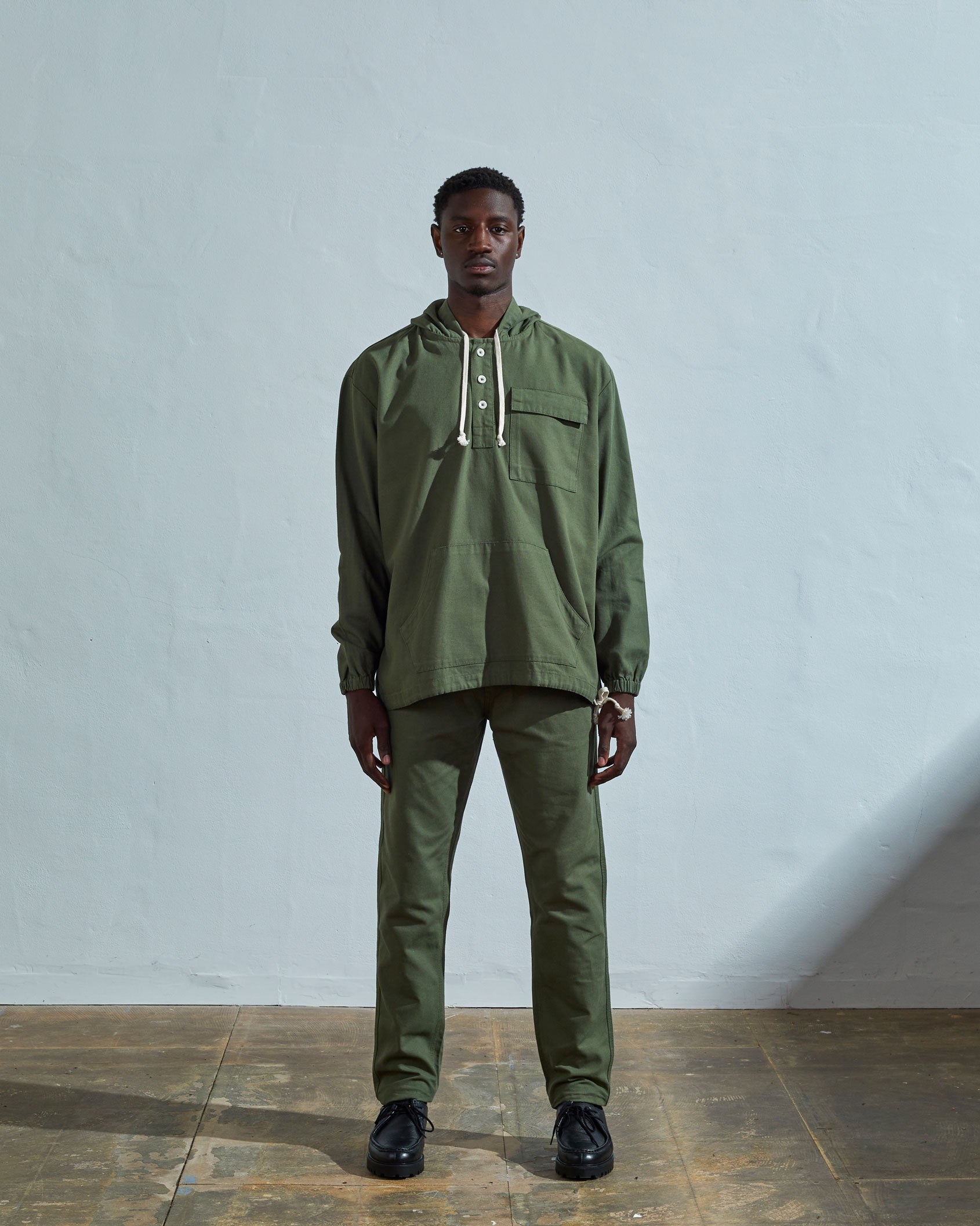 Full-length front view wearing Uskees 'coriander-green' smock, with view of corozo buttons, ribbed cuffs and drawstrings.